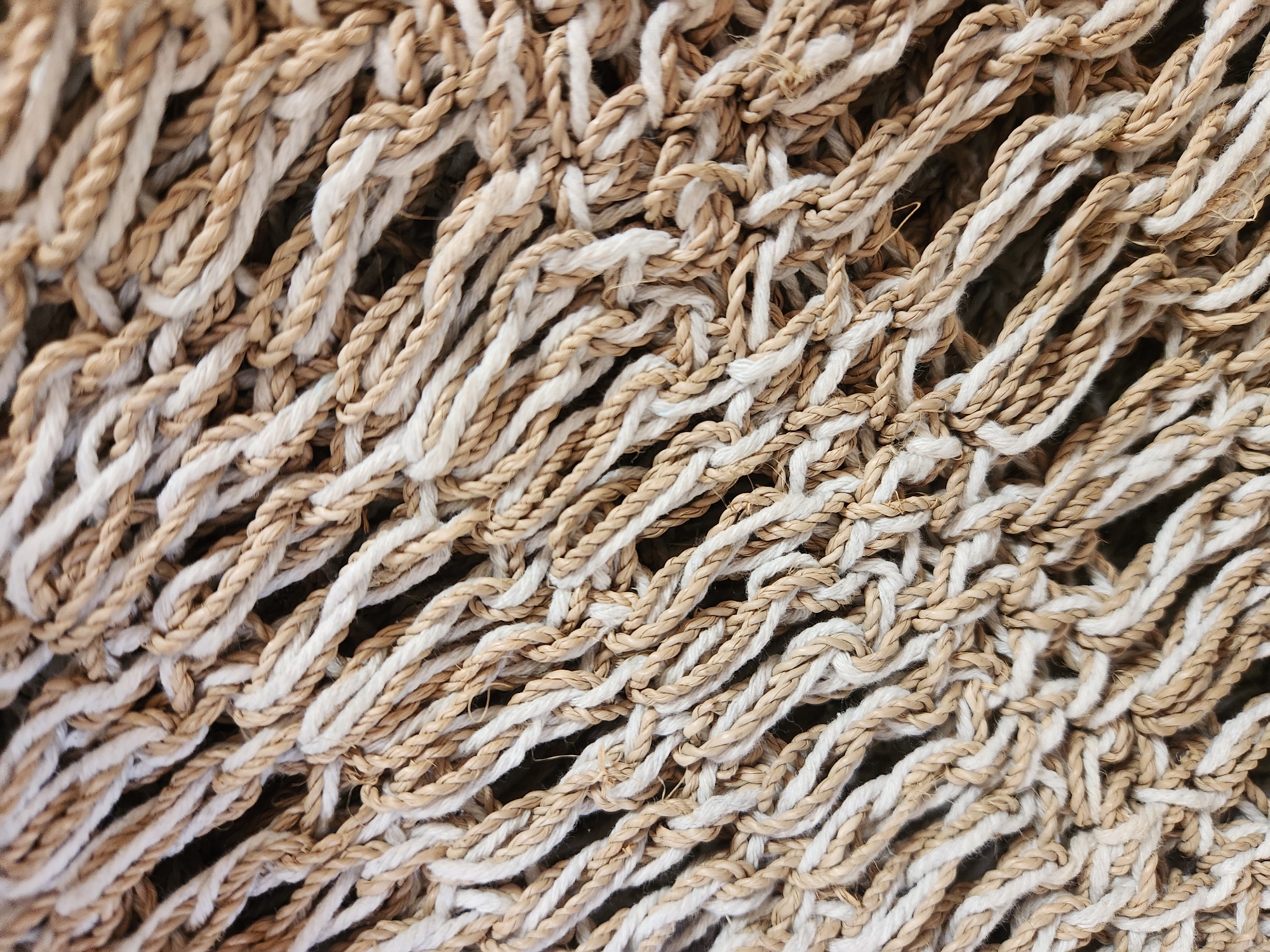 The image shows a close-up, overhead view of a tightly woven net made of thick, twisted ropes. The ropes are a mixture of off-white and natural tan colors, creating a textured, crisscrossing pattern. The material appears to be a natural fiber, possibly jute or hemp, suggesting it could be part of a hammock, fishing net, or decorative element. There is no visible indication of location within Maitland, United States of America, nor are there any people, actions, or interactions depicted. The scene is purely an abstract study of the material's texture and pattern.