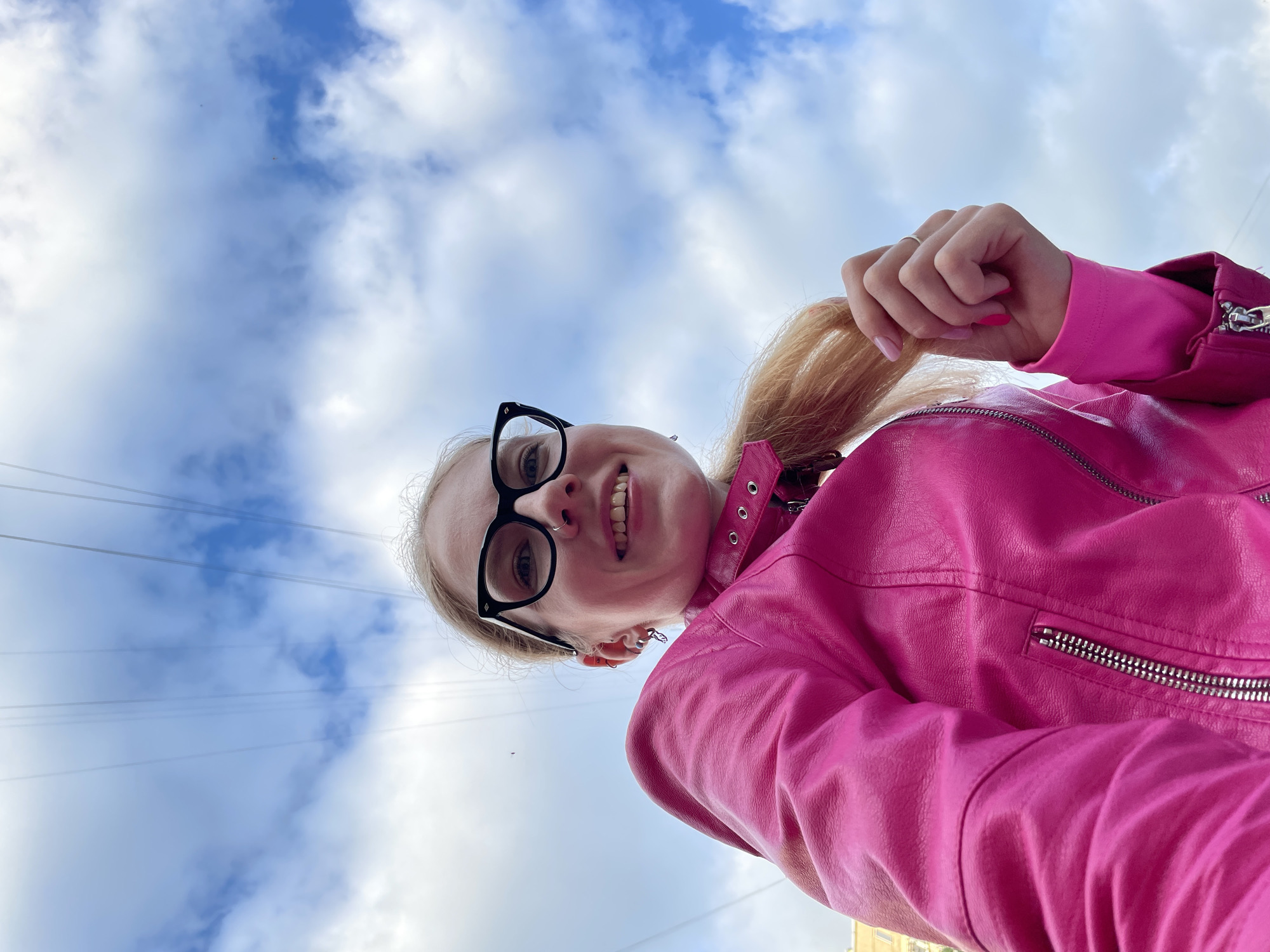 A low-angle, eye-level, outdoor, medium shot shows a woman holding her blonde ponytail. She is wearing a bright pink leather jacket with silver zippers. She has black-rimmed glasses and her mouth is open in a smile. The background is a sky with white clouds and some overhead power lines. The setting is the city of Izhevsk, Russia, likely outdoors on a street.