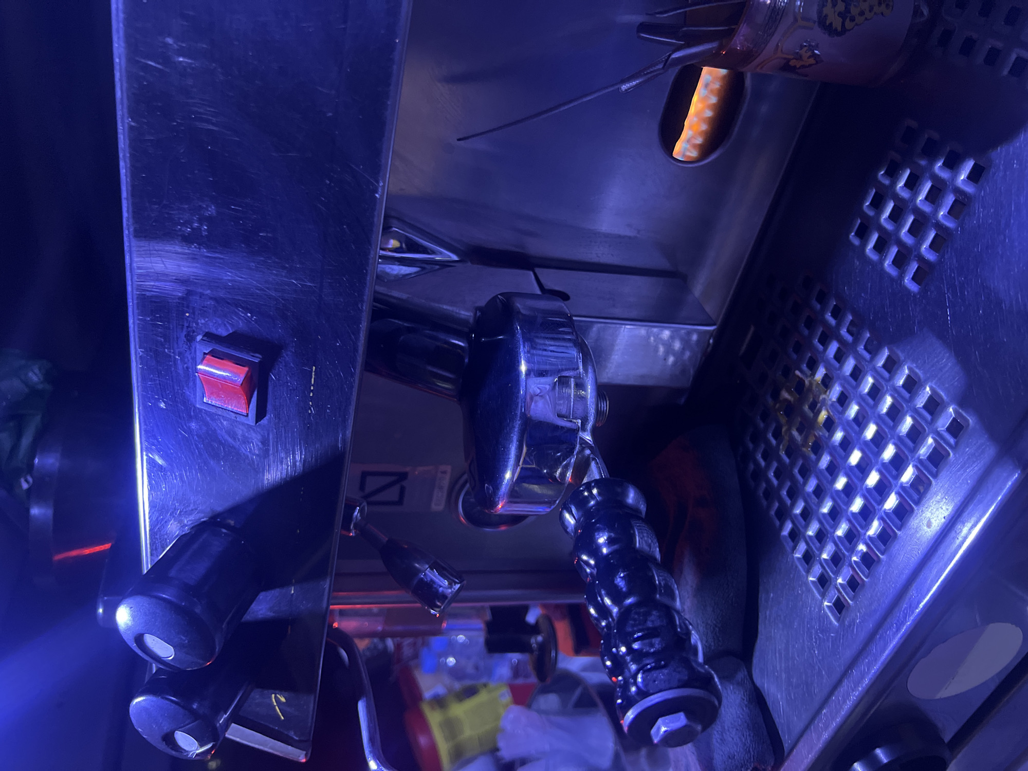 The image presents a close-up, angled view of what appears to be a commercial beverage machine, likely an espresso maker, set in a dimly lit indoor environment dominated by blue and purple atmospheric lighting. The scene suggests a night time setting.
On the left, a dark panel features a red toggle switch, below which a black handle is visible. Central to the frame are the gleaming chrome components of the machine, including a prominent ribbed, black handle, resembling a portafilter, hanging downwards. Another smaller, black handle is situated nearby.
To the right, the machine's body features a textured grate with square perforations, possibly for ventilation or as a warming surface. In the upper right corner, a section of a patterned container with an orange-yellow design is visible. Inside this container, a bright orange-yellow glow emanates, suggesting a heating element or hot coals, which could be used for specific brewing methods or a shisha service.
The background beneath the machine's main structure is blurred, revealing indistinct shapes of bottles, containers, and a yellow crate, indicating a functional, possibly cluttered, workspace. A subtle symbol, resembling a 'Z' with a diagonal line through it, is faintly visible below the main chrome assembly. No people are visible, but the setup implies an active establishment. The strong blue lighting creates a cool, atmospheric tone for this location in Martil, Morocco.