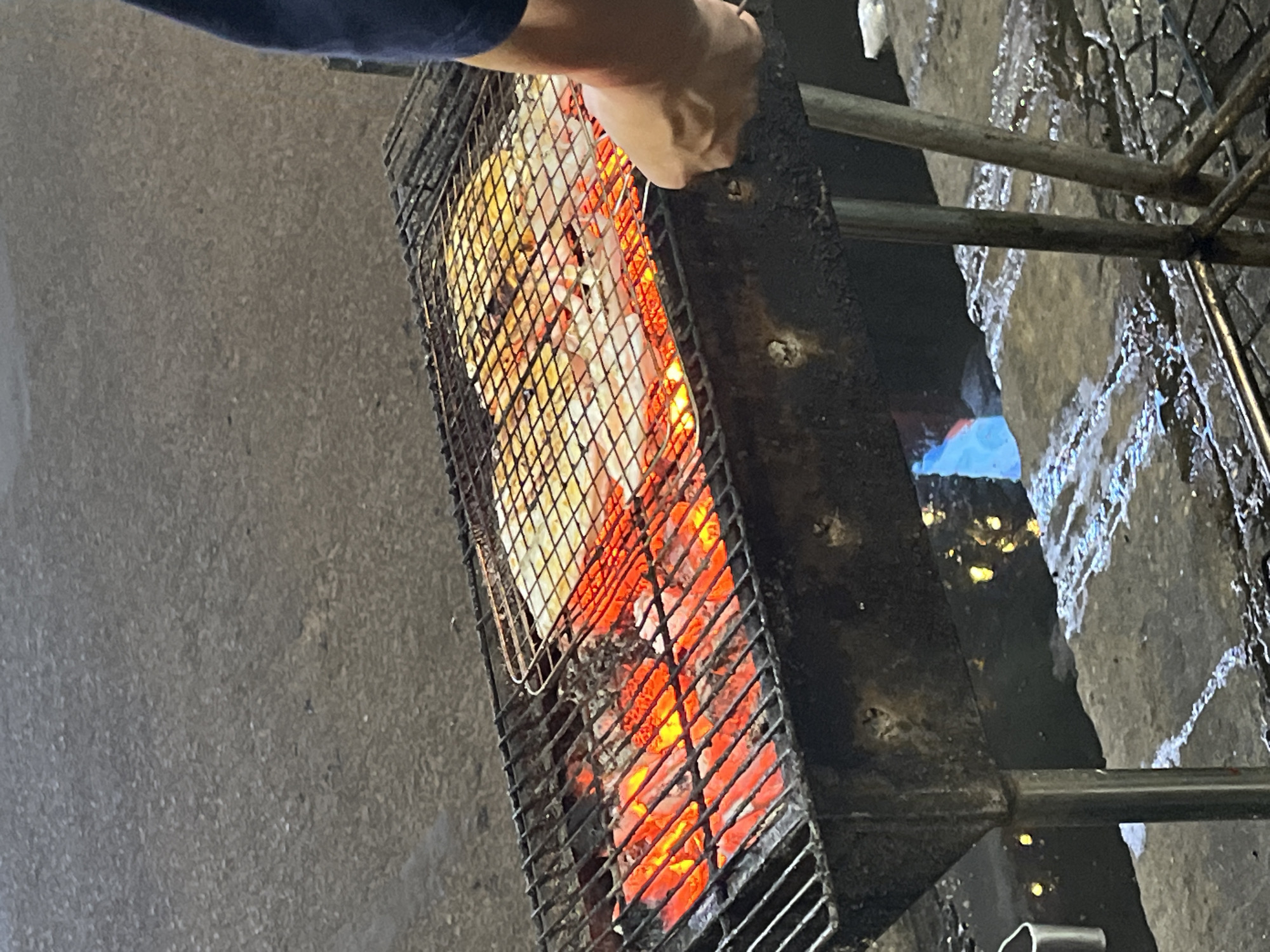 An overhead, slightly tilted shot captures a person grilling food on a charcoal barbecue grill in what appears to be an outdoor, evening setting. The primary subject is a person's right arm and hand, clad in a dark blue long-sleeved garment, holding a metal grilling basket or rack containing white-colored food, possibly seafood like squid or fish, over intensely glowing red charcoal.
The grill itself is a dark, rectangular metal structure, supported by simple metal rods. To the right of the charcoal bed, a distinct blue flame is visible, suggesting the use of a gas torch for additional heat or quick charring. The ground beneath and around the grill is dark, rough, and appears wet and uneven, possibly cobblestones or worn concrete, reflecting some light and indicating recent rain or water usage. The overall lighting is dim, emphasizing the bright orange-red glow of the coals and the blue flame. The scene evokes a street food environment, consistent with the given location context of Đà Nẵng, Vietnam. No text is visible in the image.