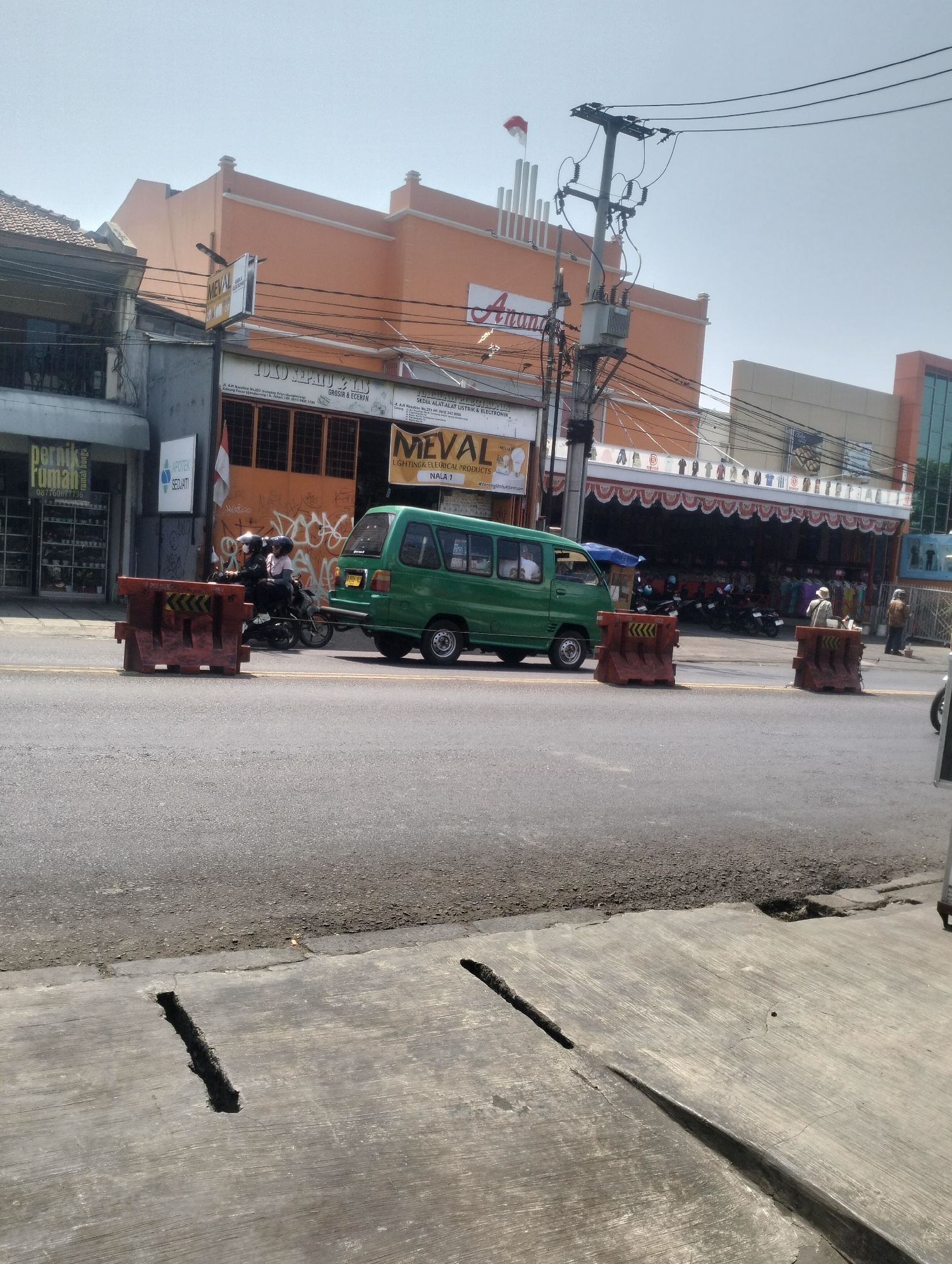 The image depicts a street scene in Kel. Pasir Jati, Indonesia, under a clear sky on a sunny day. In the foreground, a grey concrete sidewalk with a significant crack runs across it. Beyond the sidewalk is an asphalt road, bordered by yellow lines. Orange traffic barriers are placed on the road, one near the left edge and two further to the right.
A green minivan is driving on the road, partially obscured by the traffic barriers. To the left of the minivan, two people are riding a motorcycle, also partially visible behind a barrier.
On the right side of the street, buildings line the road. The buildings are of varying heights and architectural styles. The most prominent building on the left is orange with white trim and appears to be a commercial establishment, possibly a store or a mall, with a sign that reads "Anand." Another building to its left has a dark facade and a sign that says "pernik rumah" (home goods). Further to the right, another commercial building has a red and white striped awning and displays clothing for sale. Several motorcycles are parked in front of this establishment.
In the background, a utility pole with electrical wires and a transformer is visible. A red and white flag is flying on a pole on top of one of the buildings. The overall scene suggests a busy commercial area.