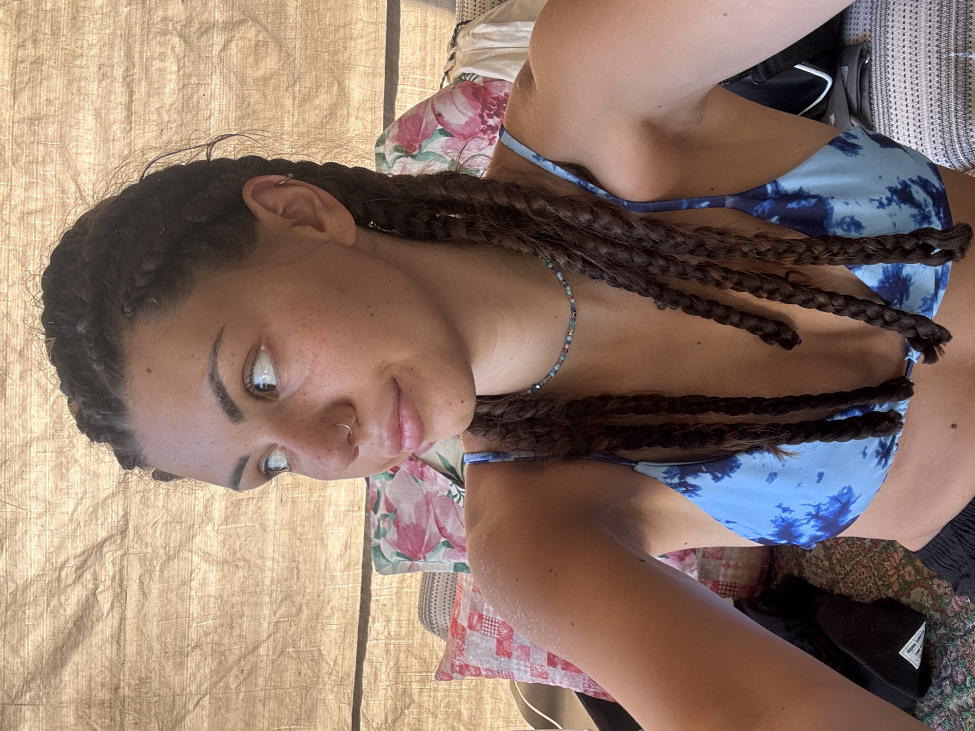A young woman with braided dark brown hair and freckles is looking to her left. She is wearing a blue tie-dye bikini top and a beaded necklace. She has a nose ring and an earring. The background consists of a textured fabric tent and decorative pillows. The setting is outdoors, likely in a camping or festival environment, with evidence of a sandy or dusty surface, consistent with a location like Belopoptsi, Bulgaria.