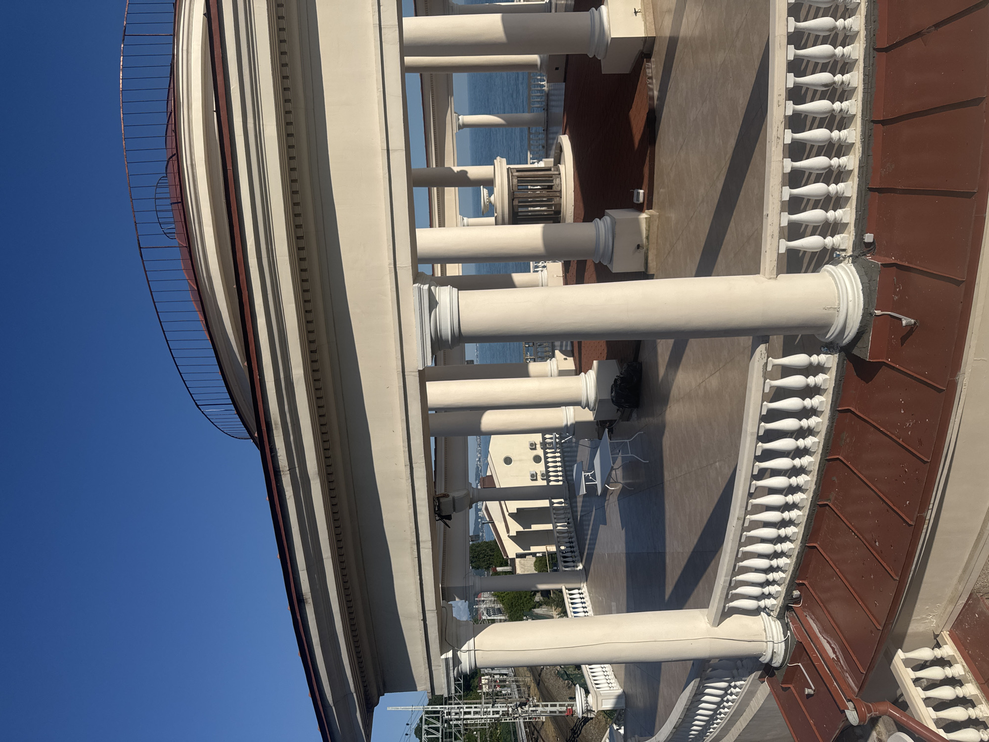 A sunlit, high-angle, medium shot captures a spacious, semi-circular colonnade overlooking the Black Sea, possibly in Sochi, Russia. The architecture features a pale cream-colored facade with a red-tiled roofline, supported by numerous large, fluted columns. A white balustrade lines the curved terrace. In the distance, the sea is visible, with a clear blue sky overhead.
On the terrace, two white tables and chairs are arranged, suggesting a seating area. Further back, a small, ornate gazebo-like structure sits in a circular planter with red tiling. The scene is uninhabited by people. Shadows from the columns and roof create a geometric pattern on the light-colored pavement. To the left, a glimpse of distant buildings and overhead power lines indicates an urban setting. The overall impression is one of architectural grandeur and a serene seaside vista.
