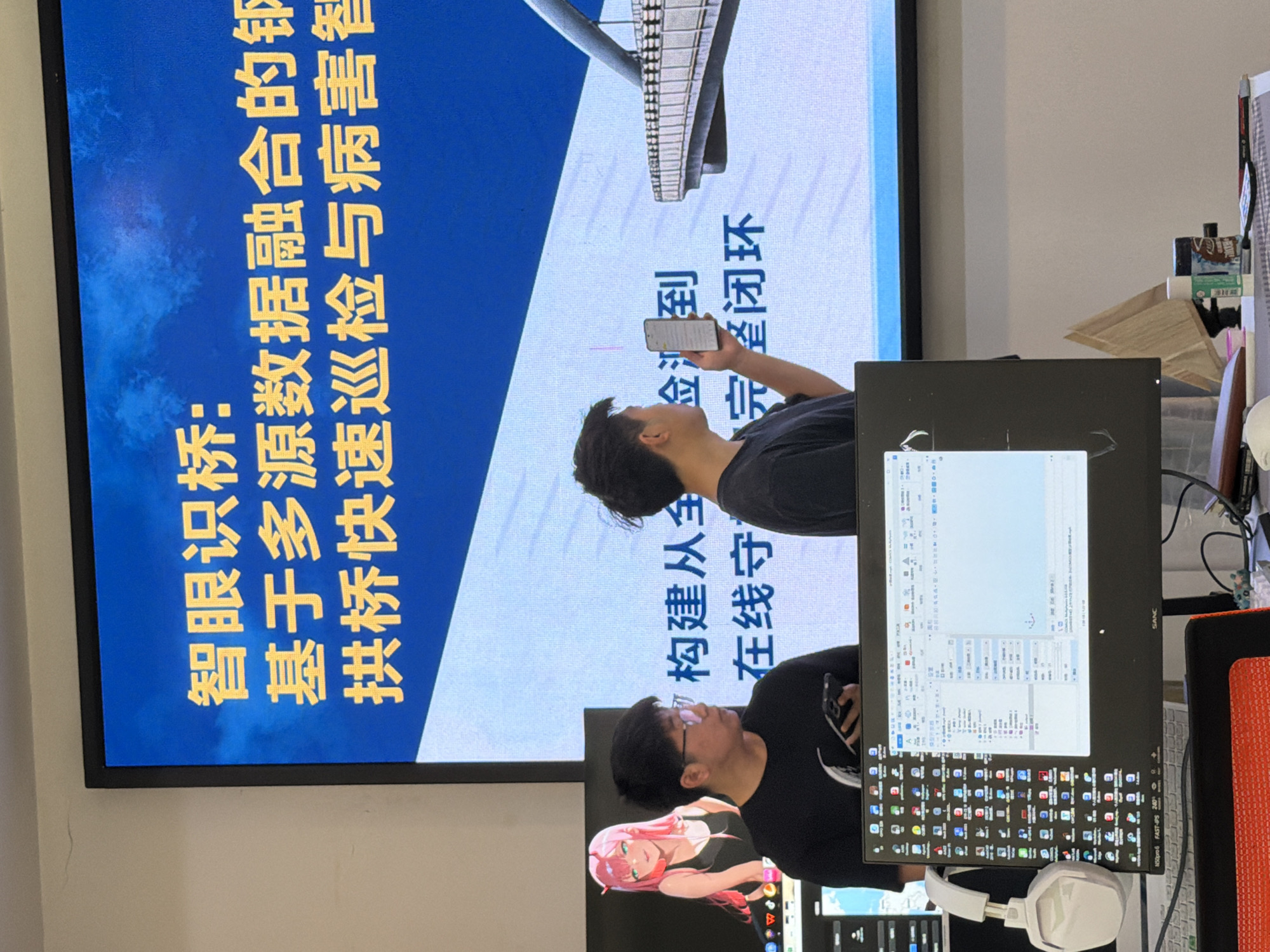 The image depicts an indoor scene featuring two individuals standing in front of a large display screen. The screen displays Chinese text and an image of a bridge. The text on the screen appears to be related to "intelligent bridge" and "multi-source data integration." One man on the left is wearing glasses and a black t-shirt, holding a smartphone in his left hand and looking towards the screen. The other man on the right, also in a black t-shirt, is holding a smartphone in his right hand and pointing at the screen. The environment includes a computer monitor displaying code and icons in the foreground, along with headphones and other office-like items. The overall setting suggests a presentation or a work environment, possibly in an office in Jiangjin, China, given the Chinese text on the screen.