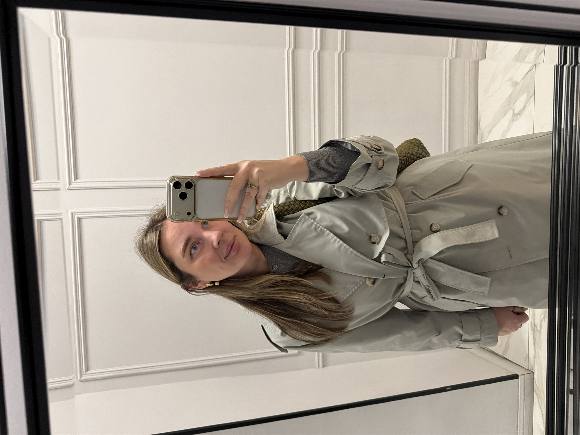 A woman is taking a selfie in a mirror. She is wearing a grey top and a light green trench coat tied at the waist. Her phone is gold. The background features white wainscoting and marble tile. The location is not discernible from the image to determine if it's in Moscow, Russia.