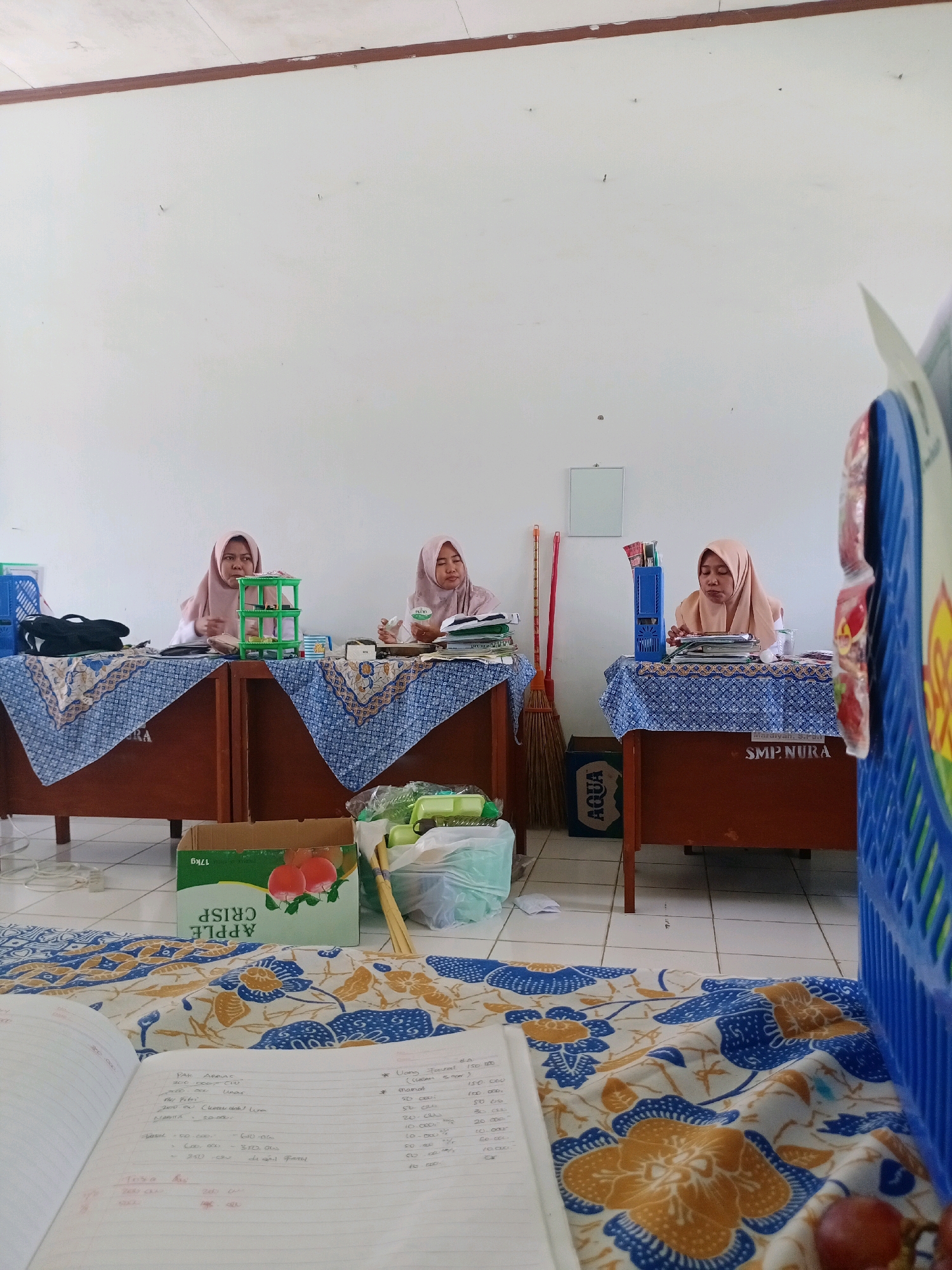 The image depicts the interior of a room, likely a classroom or office, in Tragah, Indonesia. Three women wearing hijabs are seated behind desks, appearing to be engaged in administrative or academic tasks. The room has white walls and a tiled floor. Desks are covered with blue and white patterned cloths. Various items are visible on the desks, including papers, pens, and containers. In the foreground, a notebook with handwritten notes and a patterned fabric are prominent. A blue plastic basket is positioned on the right side of the frame, and a box of "APPLE CRISP" fruit is on the floor. A broom leans against one of the desks. The overall impression is one of a functional, albeit somewhat cluttered, working environment.