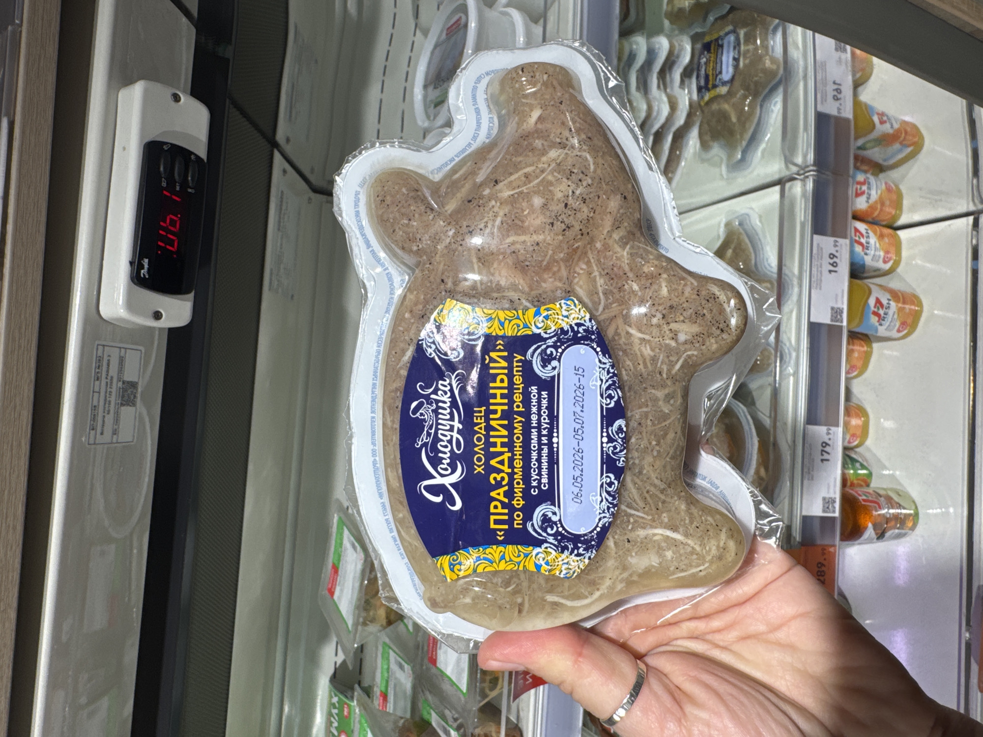 A hand holds a package of "Kholodets" or jellied meat, from a grocery store refrigerator in Moscow, Russia. The label says "Holiday Kholodets" with pork and chicken. The expiration dates are 06/05/2026 - 05/07/2026. The refrigerator temperature gauge reads 06.1 degrees Celsius. Juices are visible on the refrigerator shelf behind the Kholodets. Prices on the shelf are visible, including 169.99, 179.99, and 289.99.
