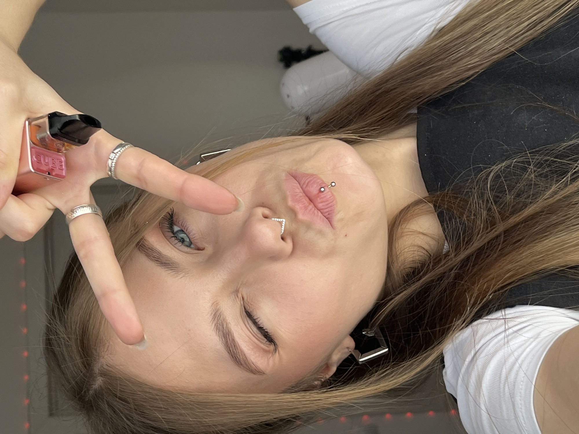 A fair-skinned young woman poses for a selfie, holding a pink "CUBE" vape pen in her right hand, wearing rings on her right hand, and displaying a peace sign. She has light brown hair, blue eyes, a nose ring, and a lip piercing. She wears a white long-sleeved shirt layered under a black top. The background is out of focus. Red lights are visible at the bottom right corner of the image. There is no indication the picture was taken in Фоки, Russia.
