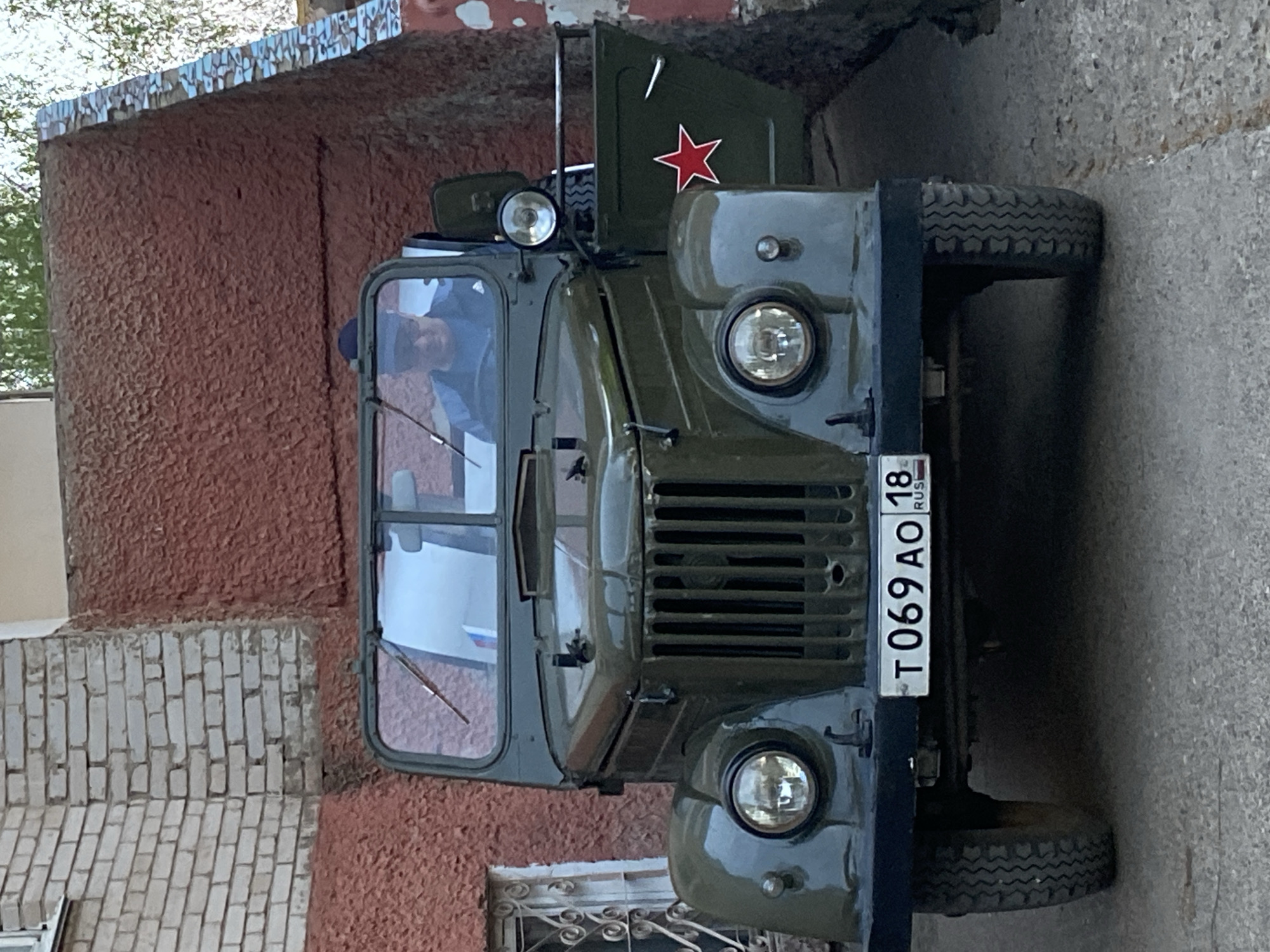 The image shows a green GAZ-66 truck parked on a street in Noviy Urengoy, Russia. The license plate reads "T 069 AO 18 RUS". The truck has a red star painted on the driver's side door. There are buildings visible in the background, including one with a red brick exterior and another with white brick. A person is visible in the driver's seat. The street appears to be paved.