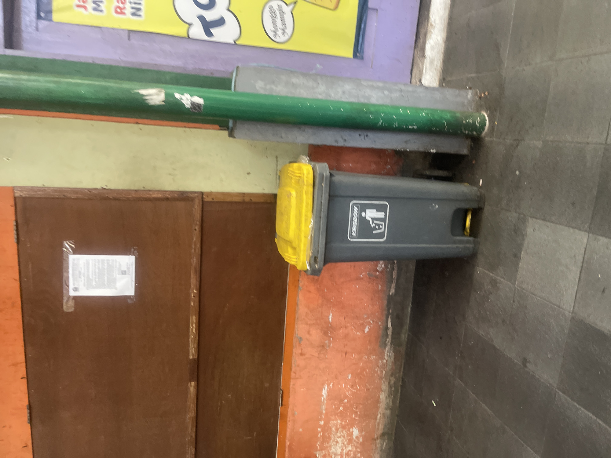 In Malang, Indonesia, a grey trash can with a yellow lid stands on a tiled floor. The trash can has a recycling symbol on its front. Behind the trash can, an orange wall is visible, and above that, a wooden structure. To the right of the trash can, a green pole stands next to a purple door. A yellow sign with text is also present on the right side of the image.