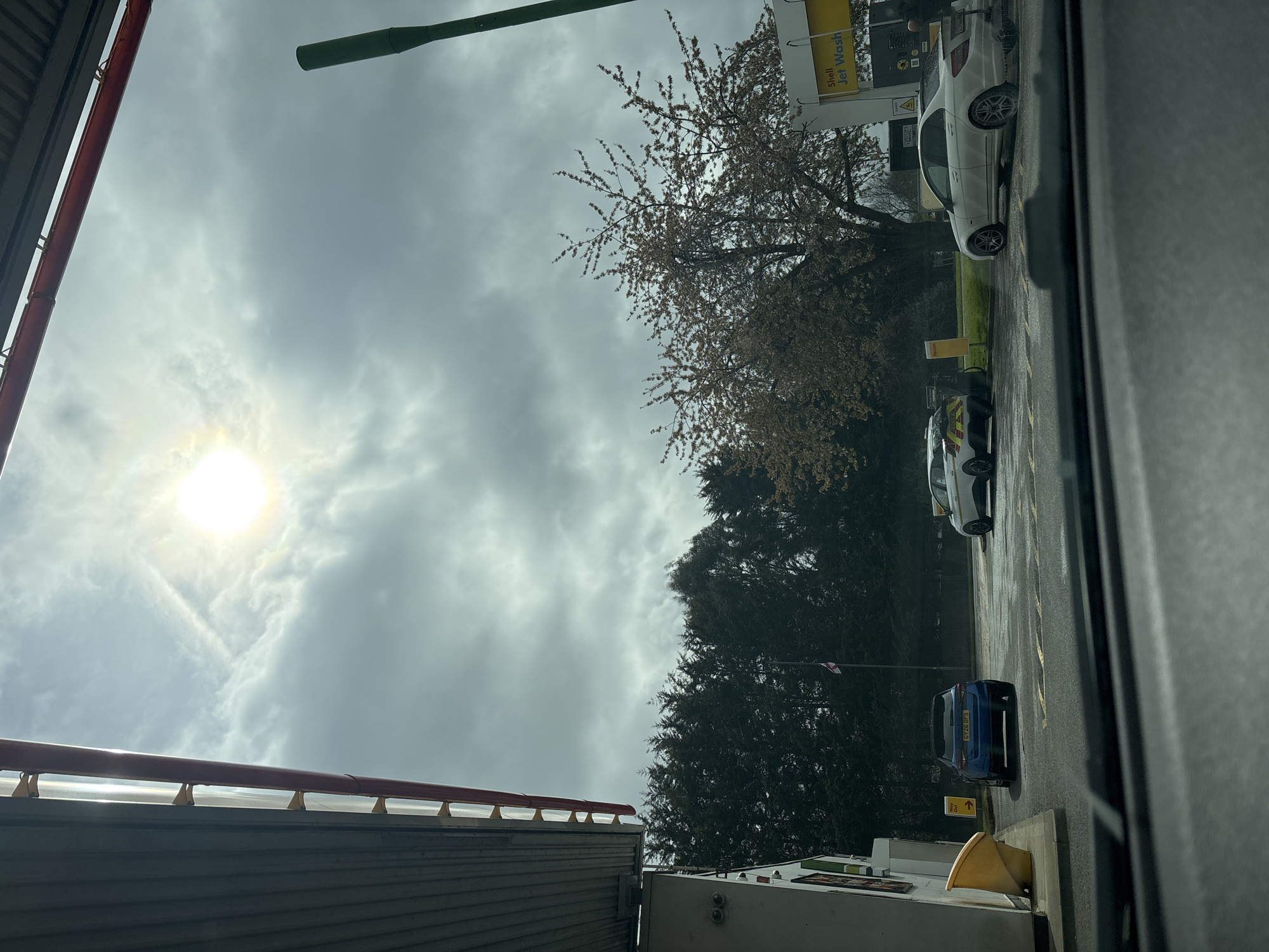 The image depicts a view from inside a vehicle, possibly a car, at a Shell petrol station in Reigate and Banstead, United Kingdom. The sun is visible through a cloudy sky. A Shell Jet Wash sign is present. Parked vehicles are visible, including a white car and a blue car with license plate "KY20 UPS". Trees and foliage are in the background. Building infrastructure elements are visible on the bottom and left side of the frame. A green beam-shaped object is visible in the top left.