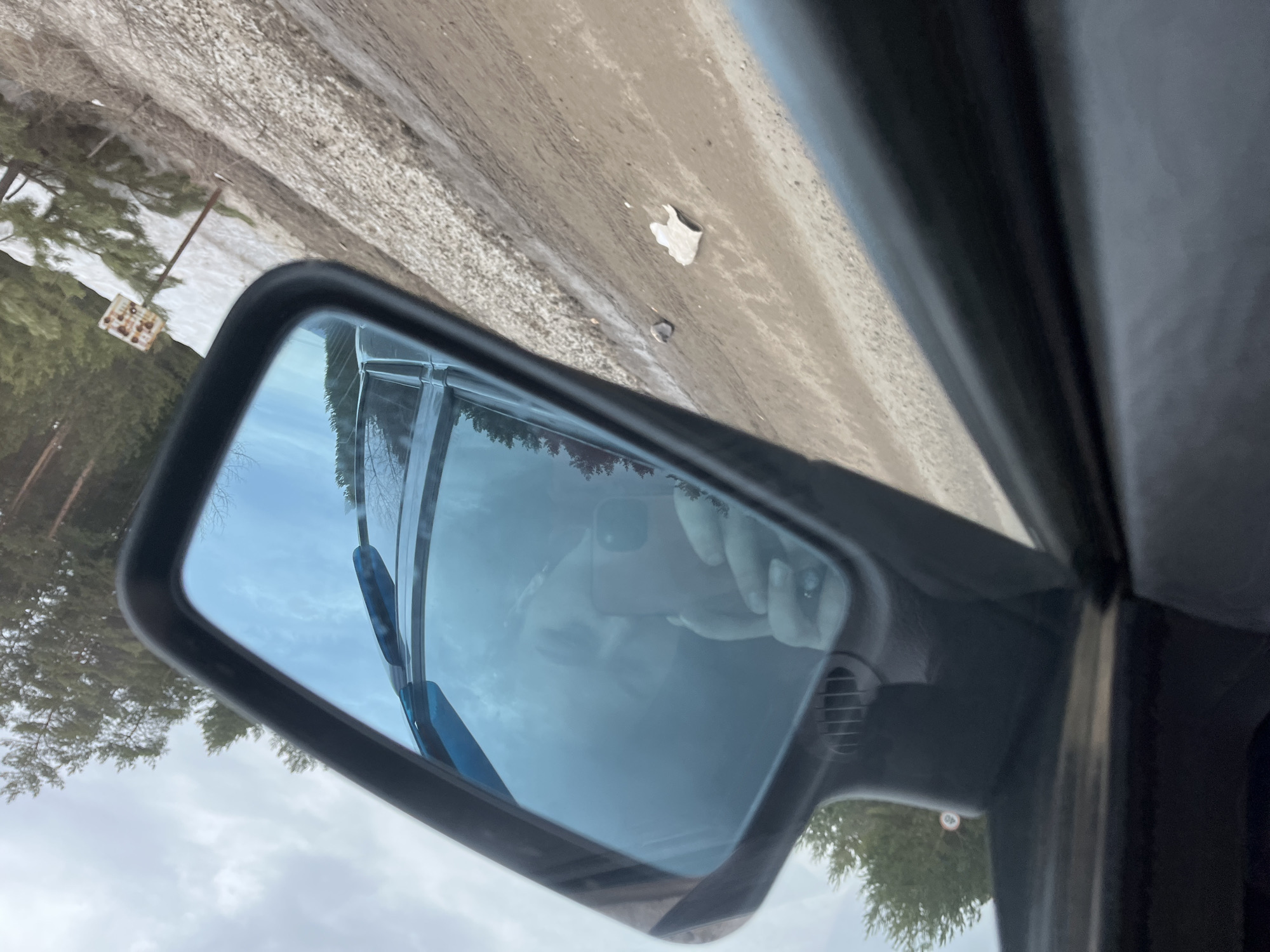 The image, taken from inside a car, shows a side mirror reflecting the surrounding environment. Reflected in the mirror are trees, the sky, and a blurred reflection of a person holding a phone, presumably taking the picture. Outside the car, the environment consists of trees, possibly a forest or park area, with patches of snow on the ground. A road runs alongside the car. A piece of white trash can be seen on the side of the road. A sign can be seen near the trees. The image was taken in the city of Chaikovsky, Russia.