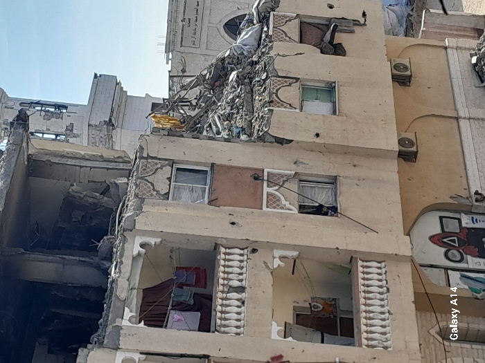 A partially destroyed building is visible. The structure reveals exposed rooms with personal items like laundry. Rubble and twisted metal protrude from the damaged sections. Adjacent to the ruined portion, a more intact building features closed windows, air conditioning units, and painted designs. The image conveys a scene of devastation and resilience.