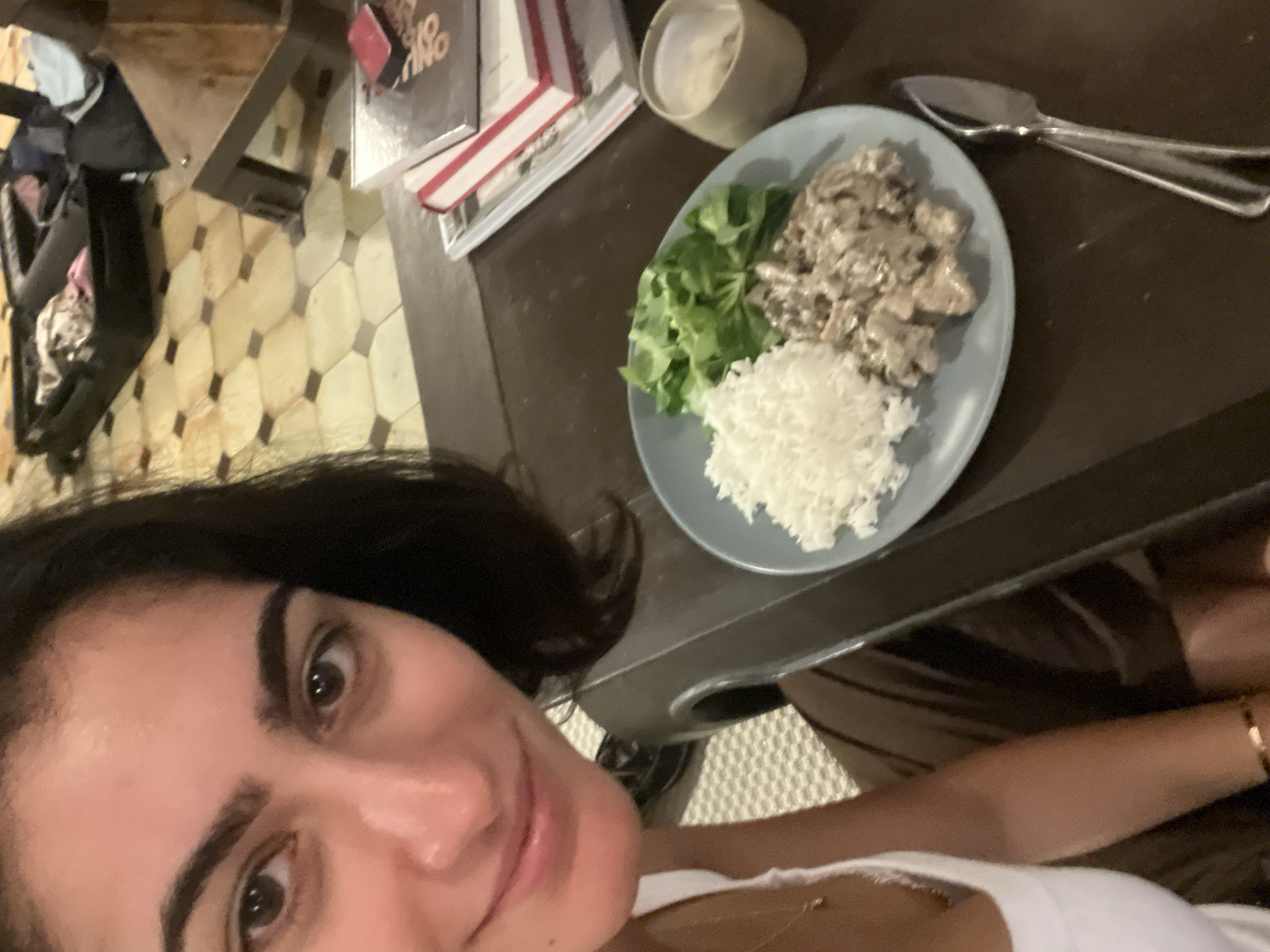 A person is taking a selfie with a plate of food. The plate is divided into three sections, one with white rice, one with green salad, and one with a creamy, meat-like mixture. Next to the plate is a set of silverware and a small ceramic bowl. The background includes a dark brown table with books or papers stacked on it, and a patterned floor. The person has dark hair and is wearing a white top. The location appears to be indoors, possibly a home or apartment. There is no definitive visual information in the image to pinpoint the location in Paris, France.