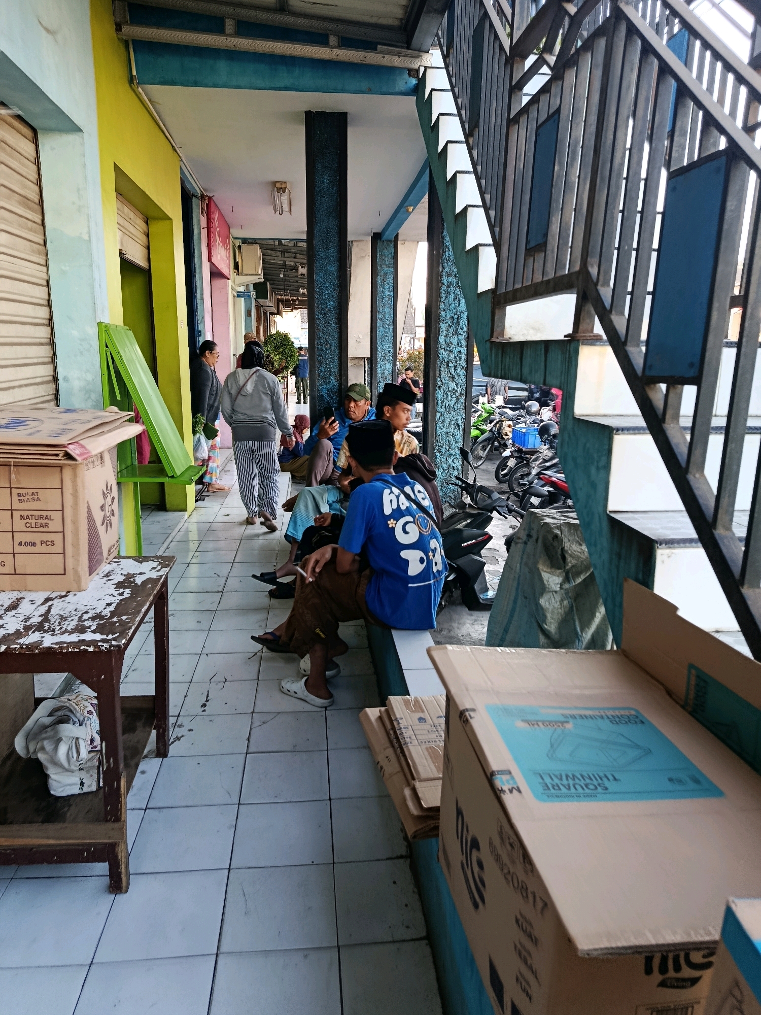 The image depicts an outdoor covered walkway, likely part of a market or shopfront, in Bululawang, Indonesia. The walkway is characterized by a tiled floor and a series of blue pillars. To the left, a row of shops with closed metallic roll-up doors is visible, painted in various bright colors including yellow and light blue. A green foldable bench is propped against one of the walls. In the foreground, a rustic wooden table with items on it, including a folded white garment, sits on the tiled floor. A large cardboard box, with text in Indonesian and English indicating it contains "SQUARE THINWALL FOOD CONTAINERS," is prominently placed on the right side of the image, obscuring part of the walkway.
Several individuals are present in the scene. In the mid-ground, a group of men are seated on a low ledge or bench under the covered walkway. One man in a blue shirt is holding a mobile phone, while another man in a brown shirt is smoking a cigarette. Further to the left, a woman in a grey hoodie and striped pants walks away from the camera, and another woman stands near her. In the background, more people are visible, and several motorcycles are parked, suggesting a busy area or transportation hub. The overall lighting suggests it is daytime, with natural light illuminating the scene. The architecture includes a metal staircase with blue panels, indicating a multi-level structure.