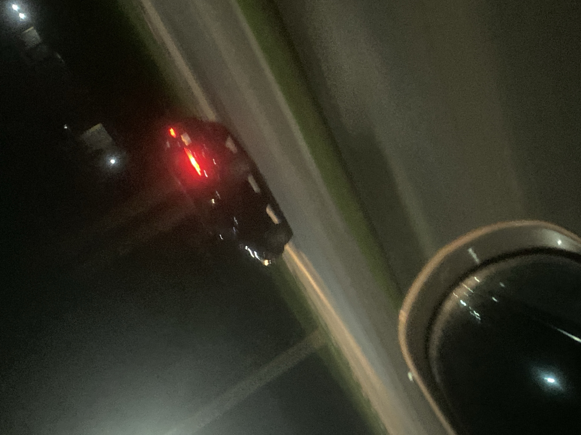 The image depicts a nighttime scene on a road in Angwan Sayawa, Nigeria. A dark-colored car is visible in motion, with its red taillights illuminated. The car appears to be a sedan. The road is paved and has a grassy shoulder. In the foreground, a portion of what seems to be a dark, rounded object, possibly a vehicle's interior or accessory, is present, reflecting some lights. The overall lighting suggests artificial streetlights and the car's own illumination, with the sky being dark.