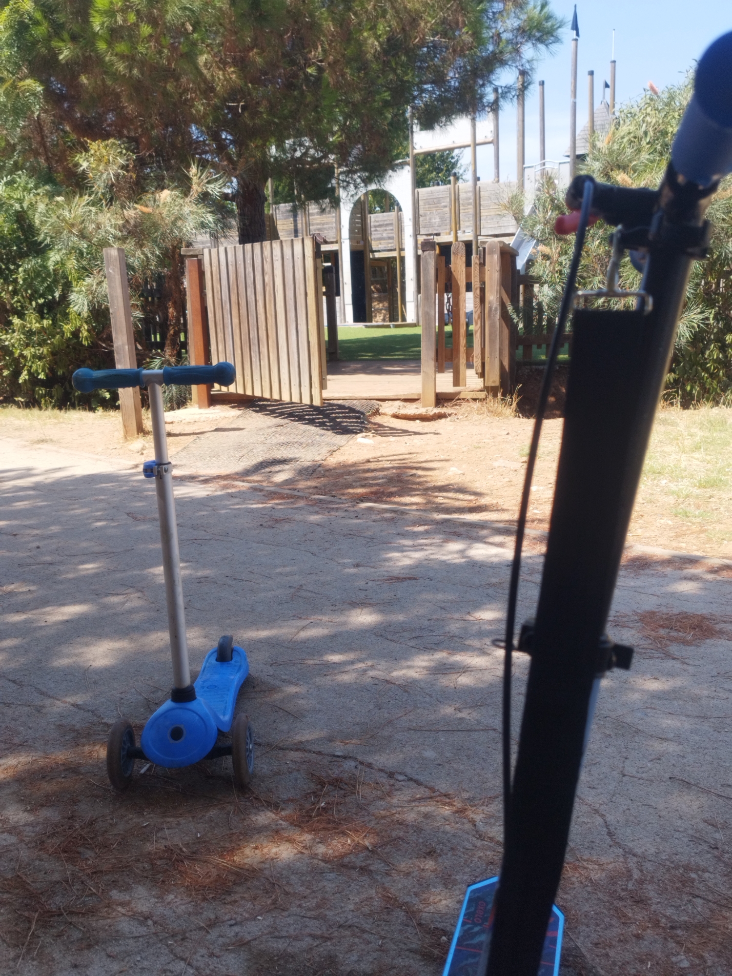 The image shows a children's playground in Antibes, France, with two scooters in the foreground. A blue three-wheeled scooter is positioned on a sandy surface to the left of the frame. In the background, a large wooden playground structure with multiple levels and slides is visible. The area is surrounded by trees and vegetation, suggesting an outdoor park setting. Sunlight casts shadows on the ground, indicating it is daytime. The image is taken from the perspective of someone standing behind a black scooter.
