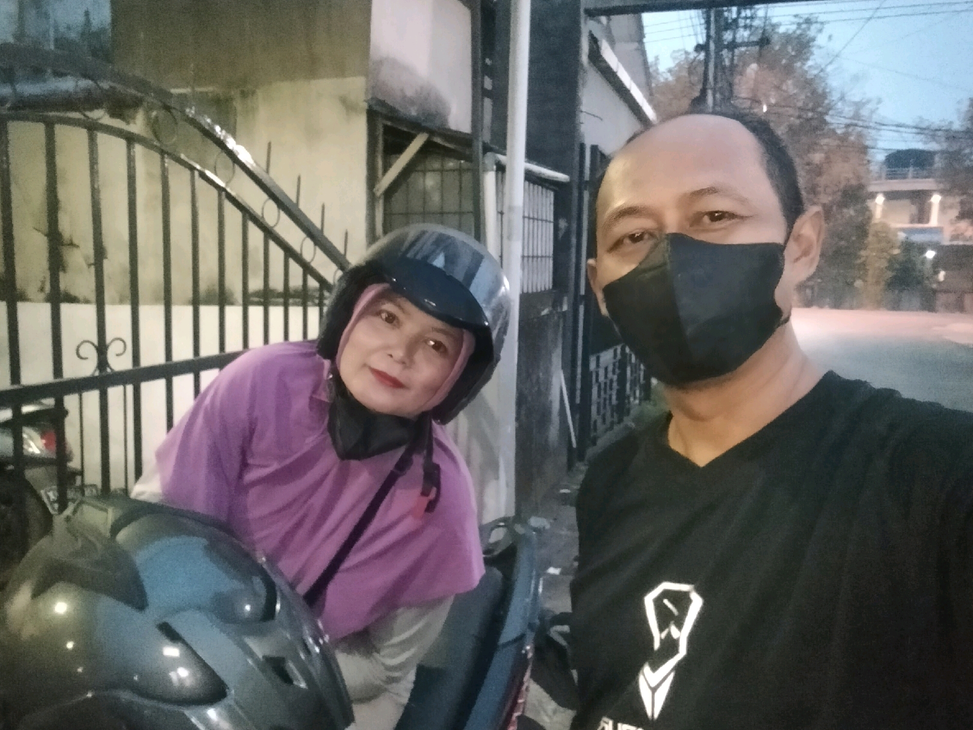 The image is a selfie taken outdoors, likely in the city of Pontianak, Indonesia. Two individuals, a man and a woman, are the primary subjects. The man, on the right, is wearing a black t-shirt with a white graphic design and a black face mask. The woman, on the left, is wearing a purple hijab and a black face mask. She is also wearing a black helmet. They appear to be sitting on a motorcycle. In the background, there is a metal fence and a building with a textured wall. Trees and a street are visible further in the distance, suggesting an urban or suburban setting. The lighting suggests it is either early morning or late evening.
