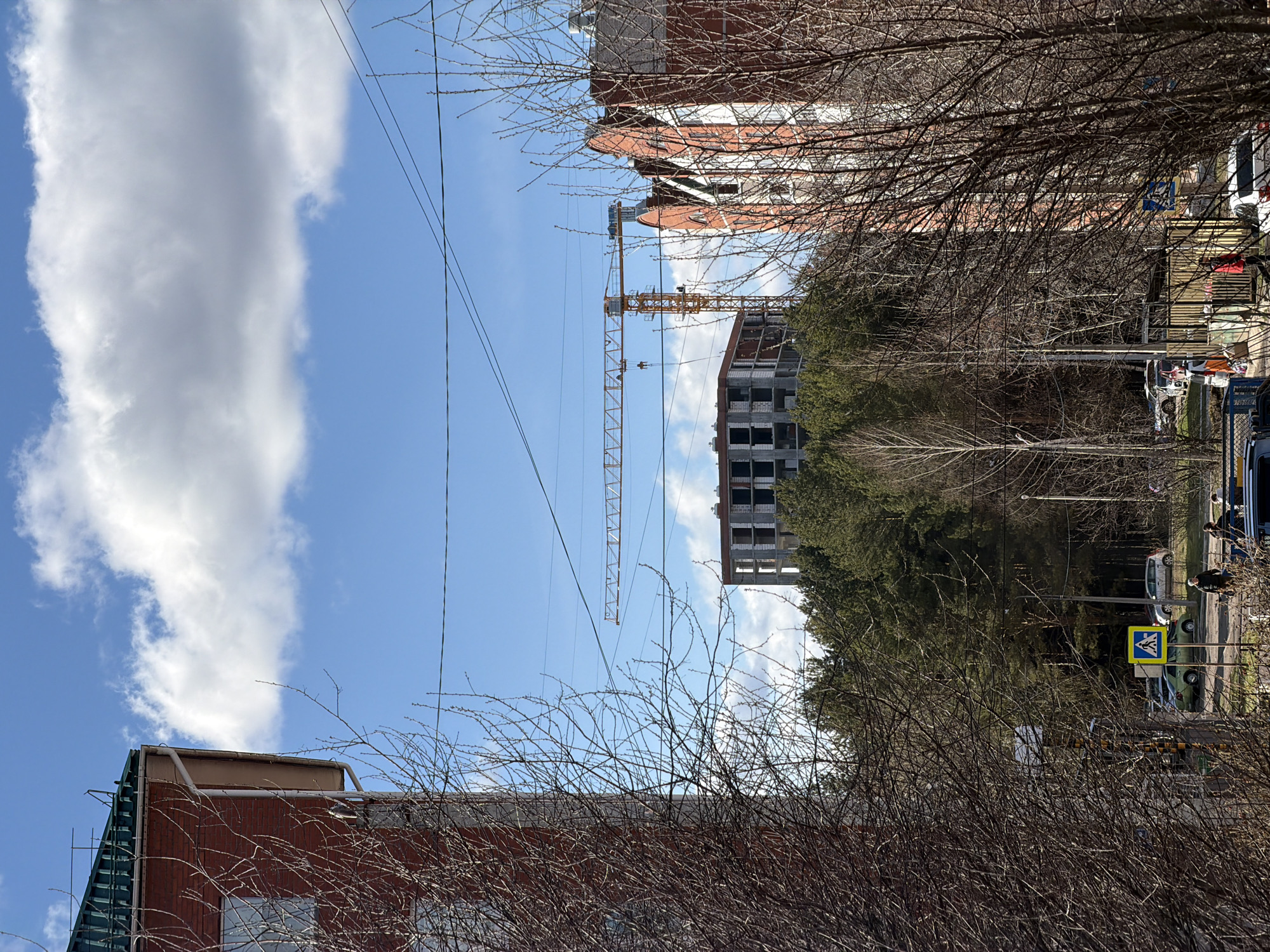 The image, taken in Chaikovsky, Russia, shows a view from ground level looking up at buildings, a construction crane, and the sky. In the foreground are bare tree branches obscuring some of the view. A red brick building with a green awning is visible on the lower left. A partially constructed building is central, and a tall yellow construction crane stands next to it. Further back, another red brick building is visible. In the background, the sky is blue with white clouds. On the right side of the image, there's some greenery, a road, vehicles and pedestrian crosswalk signs. Some people are partially visible on the right side of the image. Overhead, power lines stretch across the sky.