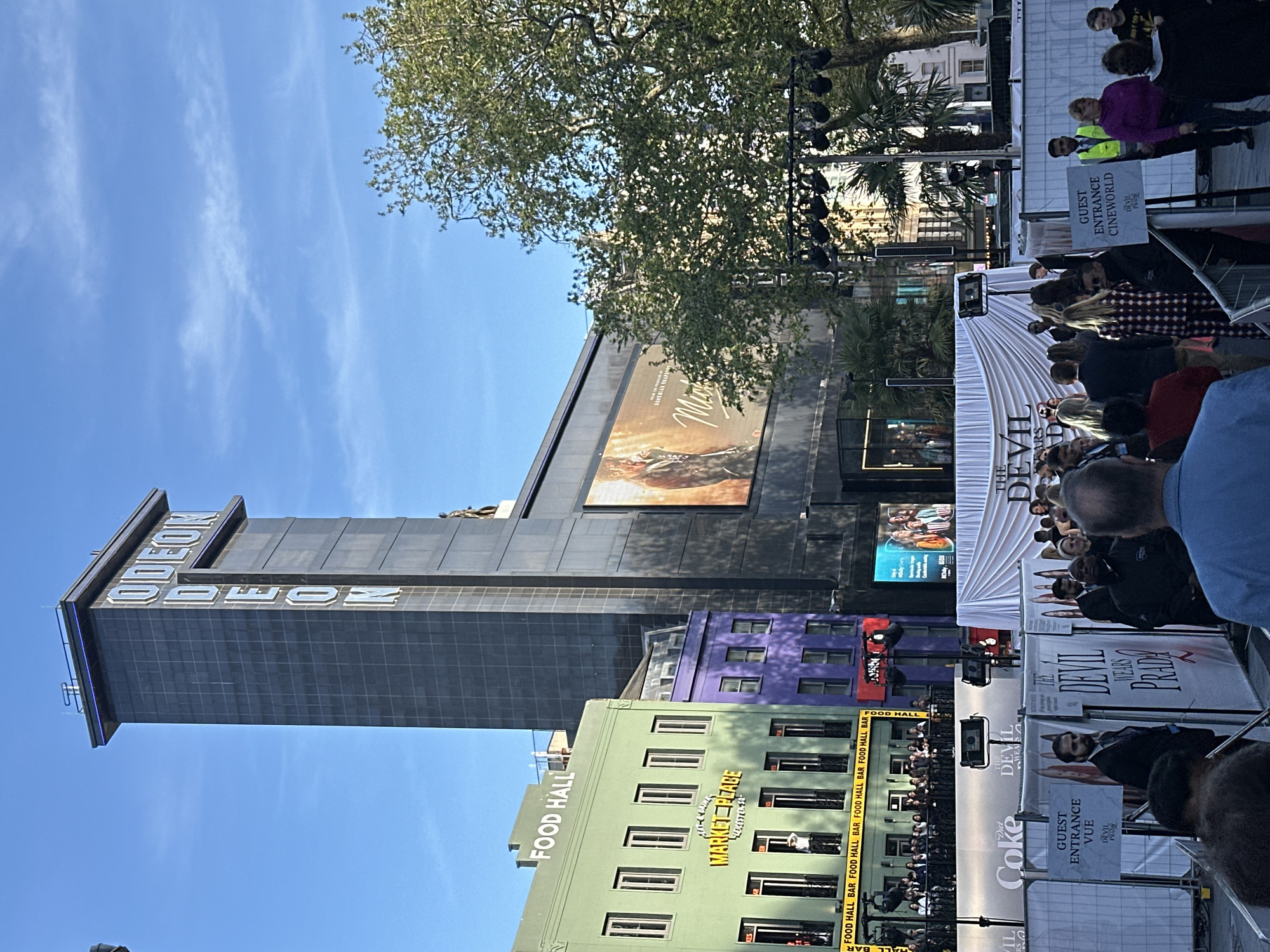 The vertical outdoor image captures a bustling public event, likely a film premiere or special screening, in a vibrant city center under a bright, clear blue sky with scattered wispy clouds. A significant crowd of people is visible, many gathered behind metal barriers, observing a white carpet event setup. Several event staff, including one wearing a yellow high-visibility vest, are present.
Prominently featured on the left is a tall, dark building adorned with the illuminated "ODEON" sign. Its facade hosts large digital billboards, one displaying the title "BOHEMIAN RHAPSODY" and another partially visible with "MAMMA MIA!". Adjacent to it stands a lime green building prominently labeled "FOOD HALL" and "MARKET PLACE," with "FOOD HALL BAR" repeated across its lower levels. A purple building is situated between the green building and the main cinema structure.
The event area itself is characterized by white banners and signs prominently featuring "THE DEVIL WEARS PRADA." Directional signage indicates "GUEST ENTRANCE CINEWORLD" and "GUEST ENTRANCE VUE," suggesting organized access for cinema attendees. Additional branding for "Diet Coke" is visible on barriers. Trees, including lush green foliage from a large tree and several palm trees, are interspersed within the urban landscape. The scene, bathed in bright daylight and featuring iconic cinema brands, is indicative of a major city location, specifically London, United Kingdom.