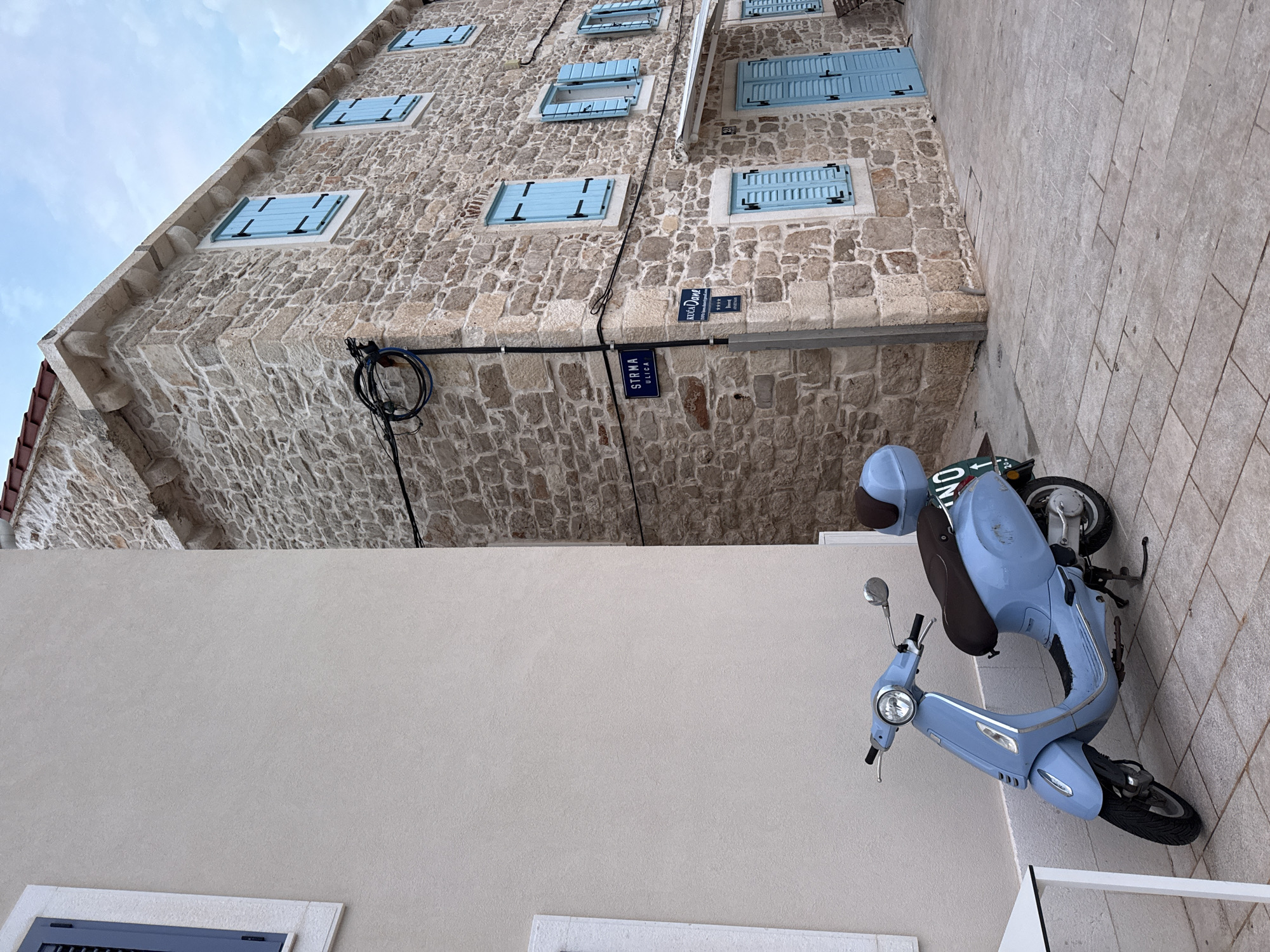 A light blue Vespa scooter is parked on a paved sidewalk in front of a stone building with blue shutters. The building is located on a narrow street in Primošten, Croatia. The street is paved with light gray stones. There is a white building on the left side of the image. The sky is cloudy.