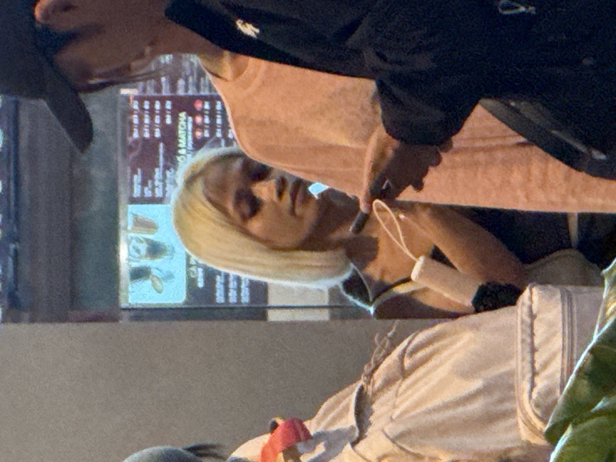 In Bắc Cam Ranh, Vietnam, a blonde-haired individual with light skin is seen in a crowd, looking downwards at their phone. The person wears a dark top and holds a white object in their right hand. To their right, another individual in a peach-colored t-shirt appears to be looking at the phone as well. Further to the right, a person wearing a black baseball cap and a dark jacket is partially visible, also looking down. Behind the blonde individual, a digital menu board displays text and images, likely indicating food and drink options. In the foreground on the left, a beige backpack with a zipped pocket is visible. The lighting suggests an indoor setting, possibly a cafe or restaurant.