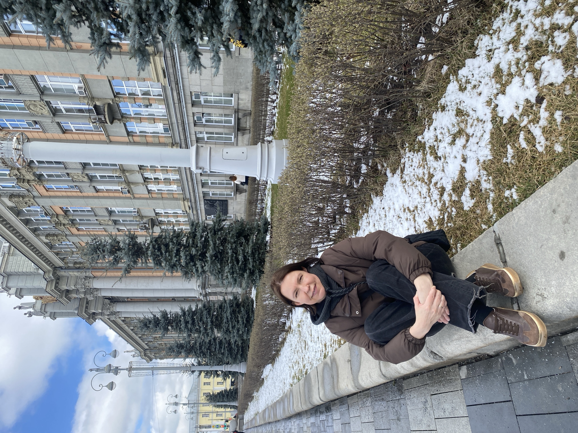 A woman is sitting on a concrete ledge in an urban environment. She is wearing a brown puffer jacket, black jeans, and brown shoes. She is hugging her knees. The ledge overlooks a grassy area with patchy snow. Behind the grassy area is a building with a classical facade, featuring numerous windows and ornate columns. The sky is visible in the lower left corner, showing some blue and white clouds. Based on the architectural style and location, this is likely in Yekaterinburg, Russia.