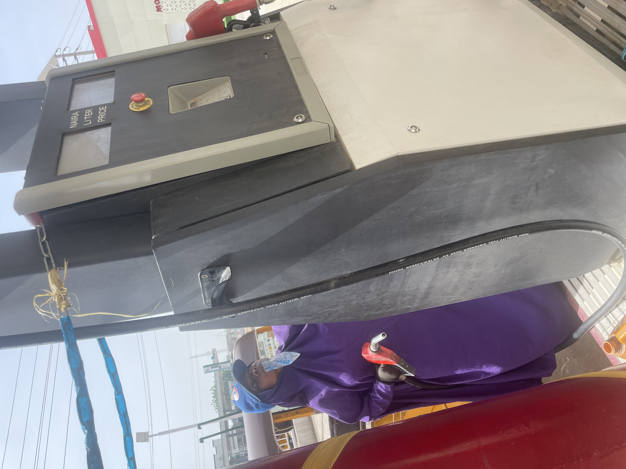 A gas station in Unknown, Unknown is depicted from a low angle, with a person in a purple robe and gloves holding a red gas pump nozzle. The nozzle is connected to a black hose extending from a gas pump. The pump displays areas for Naira, Liter, and Price indicators, alongside an emergency stop button and a number pad. A red gas tank is visible below. The surrounding environment shows other buildings and the sky.