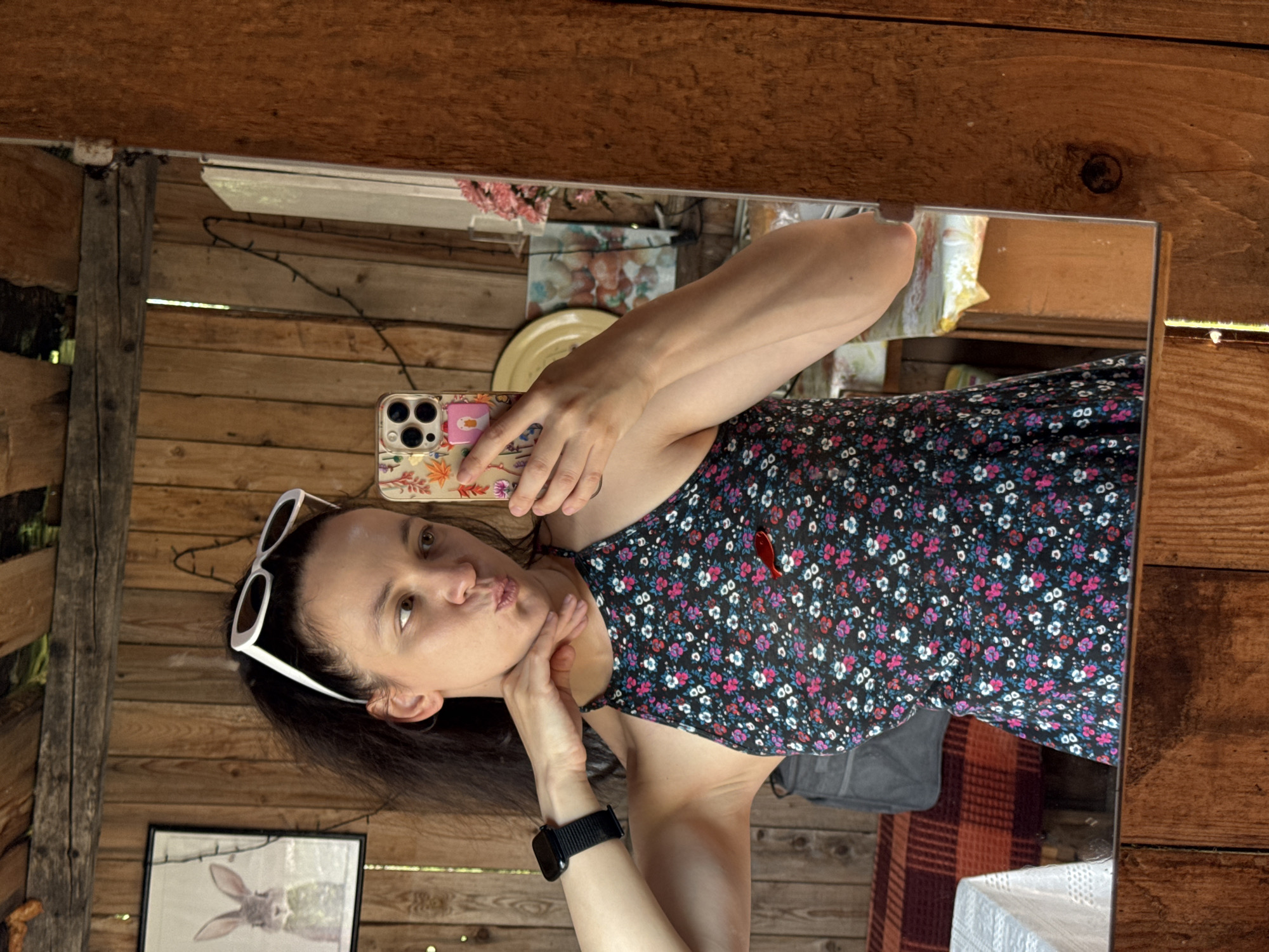 A woman with dark hair pulled back, wearing white sunglasses on her head and a floral print dress, is taking a selfie in a mirror. She is holding a phone with a colorful case in her right hand and touching her chin with her left hand, making a pouty face. She is wearing a black watch on her left wrist. The background appears to be an interior made of wooden planks, possibly a rustic cabin or shed. A framed picture of a rabbit is visible on the wall to her left. A gray bag is partially visible behind her. The scene is well-lit, with natural light contributing to the overall ambiance. The location in the city of Zasechnyi, Russia is not specified in the image.