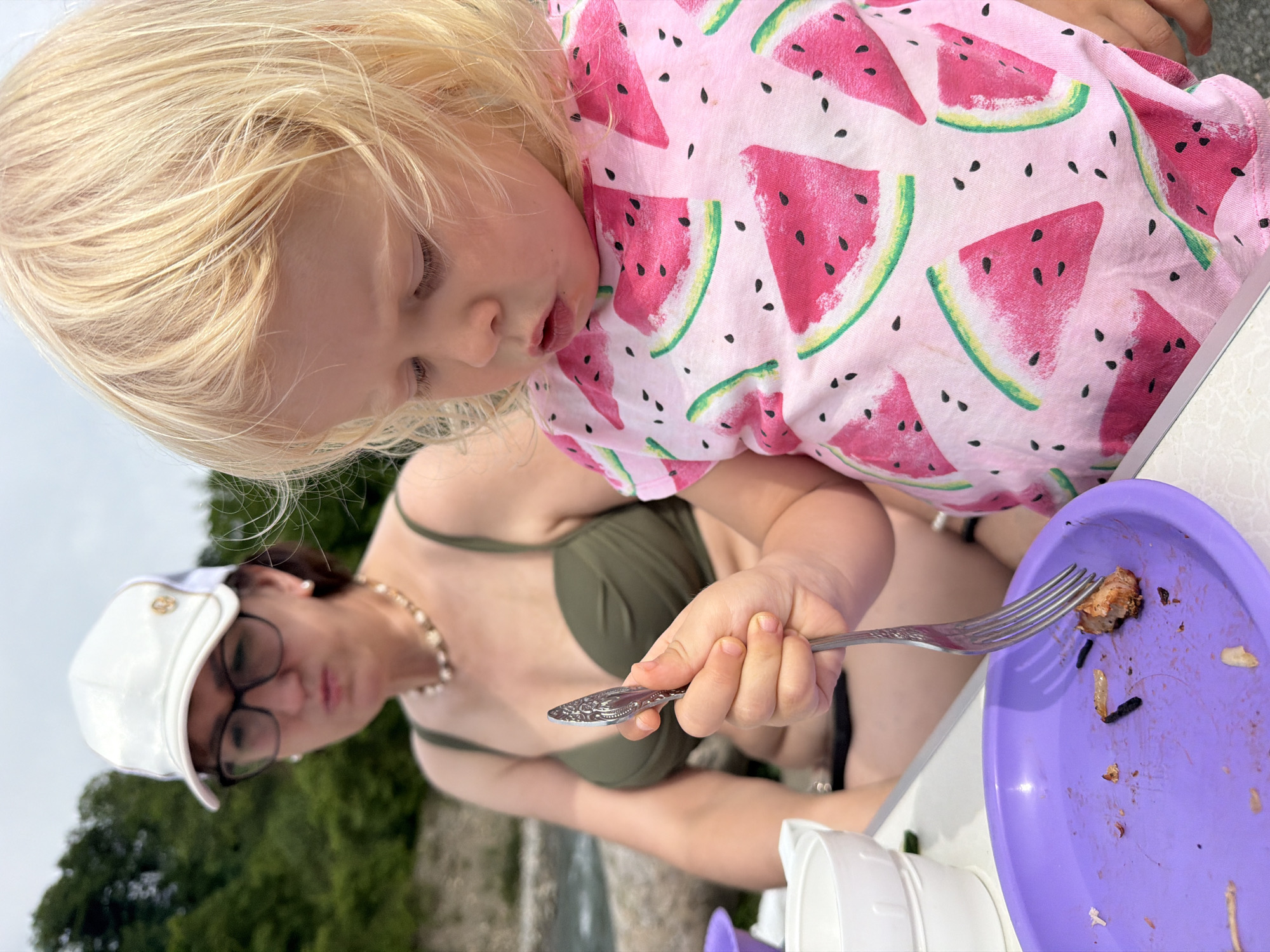 In a medium shot, a young child with blonde hair is focused on a purple plastic plate with a fork. The child is wearing a pink t-shirt adorned with watermelon slices. The child's face is partially obscured as they lean over the plate, but their expression appears concentrated. A woman wearing glasses and a white cap with a gold emblem is visible in the background, to the left of the child. She is also wearing a green bikini top and a pearl necklace.
The scene appears to be outdoors, with a backdrop of blurred green foliage and what seems to be a body of water. The lighting suggests it could be daytime, possibly overcast. The overall atmosphere is casual and relaxed, likely a mealtime setting. The child is actively engaged with the food on the plate, using the fork to pick up a piece of food. The plate has remnants of food, indicating a meal is in progress or has recently concluded.
The presence of a beach umbrella or similar structure is hinted at by the white plastic cup on the right side of the frame. The image was taken in Sochi, Russia, suggesting a possible vacation or outdoor recreational activity. No visible text is present in the image.