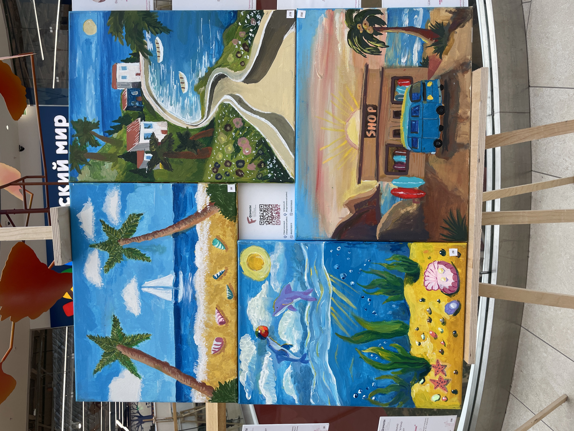 Four paintings are displayed on wooden easels in what appears to be an indoor public space. The paintings depict various beach and seaside scenes. The background of the image shows signage with Cyrillic text, one of which reads "СКИЙ МИР" (SKY WORLD). A QR code is visible on a white label on the second painting from the top left, beneath it is text which reads "ФЭМИЛИ" (FAMILY). Below this, there is text in Cyrillic characters that includes "Екатеринбург" (Yekaterinburg), suggesting a connection to that city. Given the presence of Cyrillic text and Russian place names in the visible information, the location is likely in Russia. The specific location of Perm, Russia, cannot be definitively determined from the visual information provided.
The painting on the top left shows a beach with two palm trees, a sailboat on the ocean, and shells scattered on the sand. The top right painting depicts a coastal village with houses, a winding road, and boats in the water. The bottom left painting shows an underwater scene with kelp, starfish, seashells, and two dolphins playing with a ball. The bottom right painting features a beach shop with surfboards, a blue van, and a palm tree against a sunset sky.