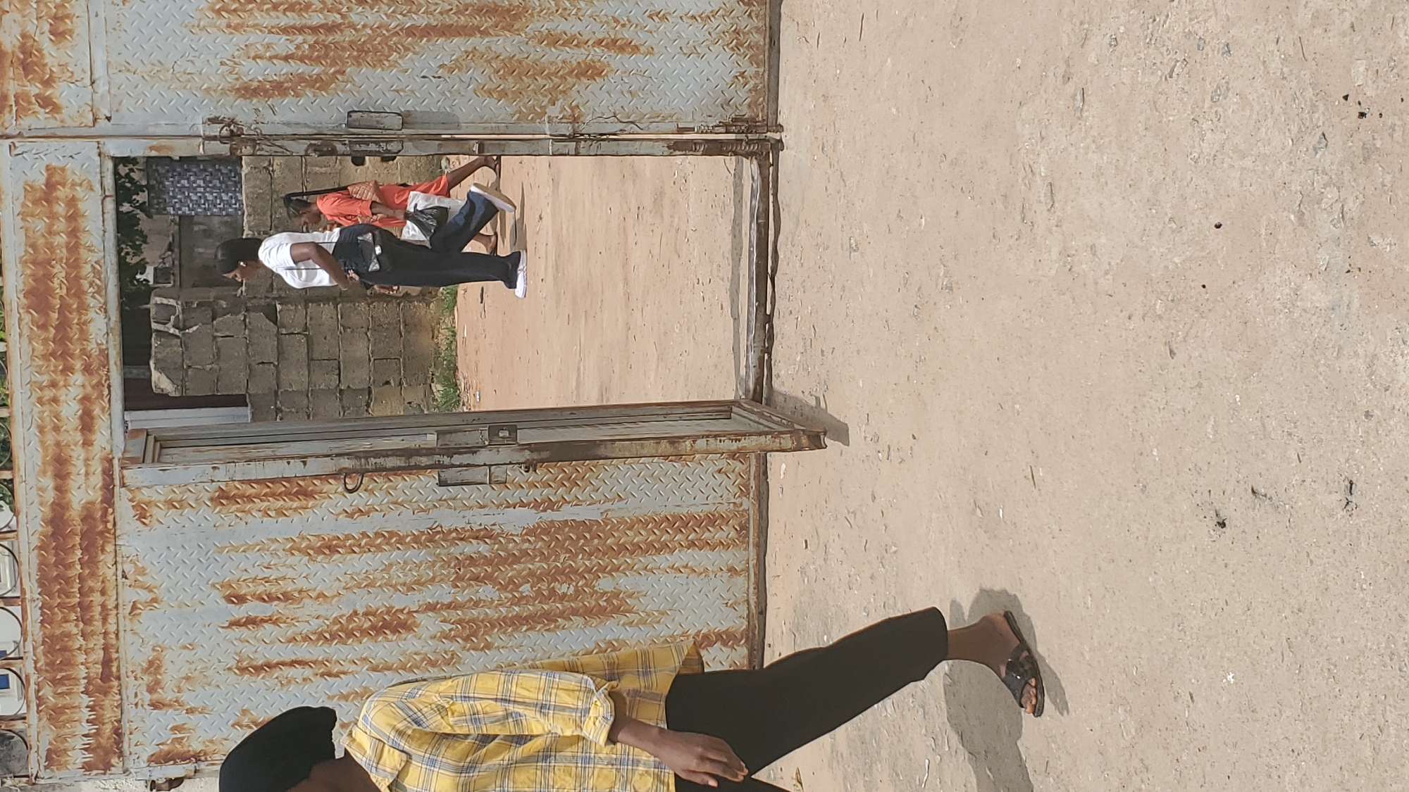 In Jalingo, Nigeria, a rough, rusty metal gate with diamond plate texture is partially open, revealing a pathway beyond. To the left, a person wearing a yellow and black plaid shirt and dark pants walks away from the camera, their feet shod in dark sandals. Through the gate, two individuals are walking towards the viewer. The first, on the left, is a woman in a white t-shirt and dark pants, carrying a black purse and a white plastic bag. To her right and slightly behind her, a child in an orange outfit is also walking. The ground appears to be made of dirt and gravel, with some debris scattered across it. The background consists of rough brick structures.