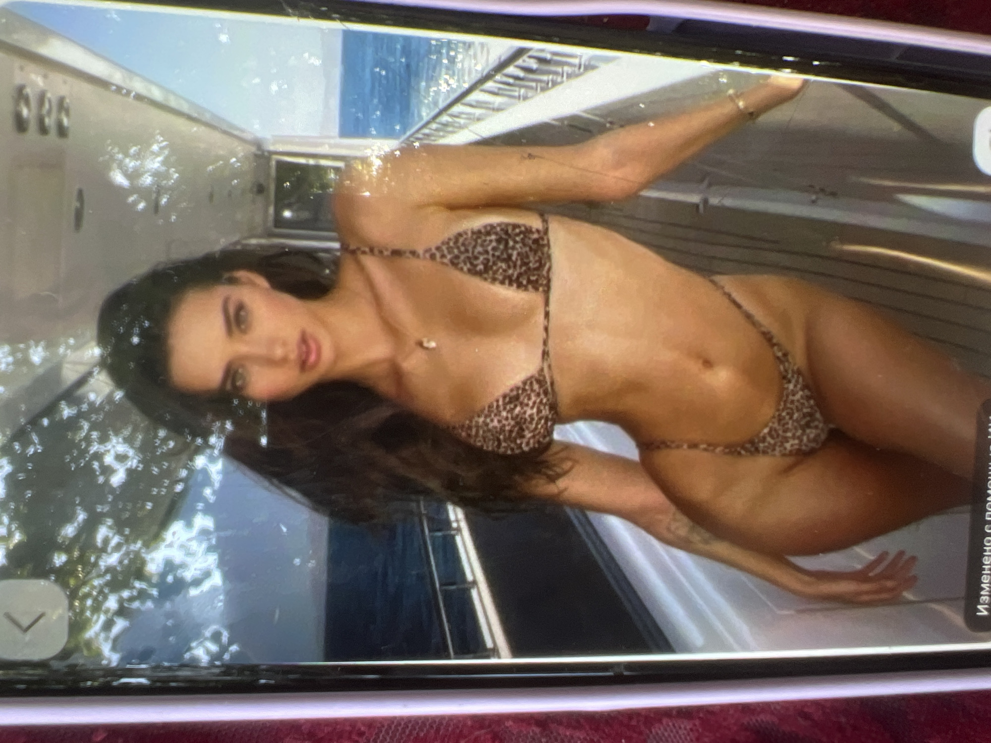 A young woman with long, dark hair and green eyes is posing on the deck of a yacht. She is wearing a leopard-print bikini and has a necklace with a small pendant. The yacht is at sea, with the ocean visible in the background. The lighting suggests it is daytime. The image is a photograph taken on a phone screen.