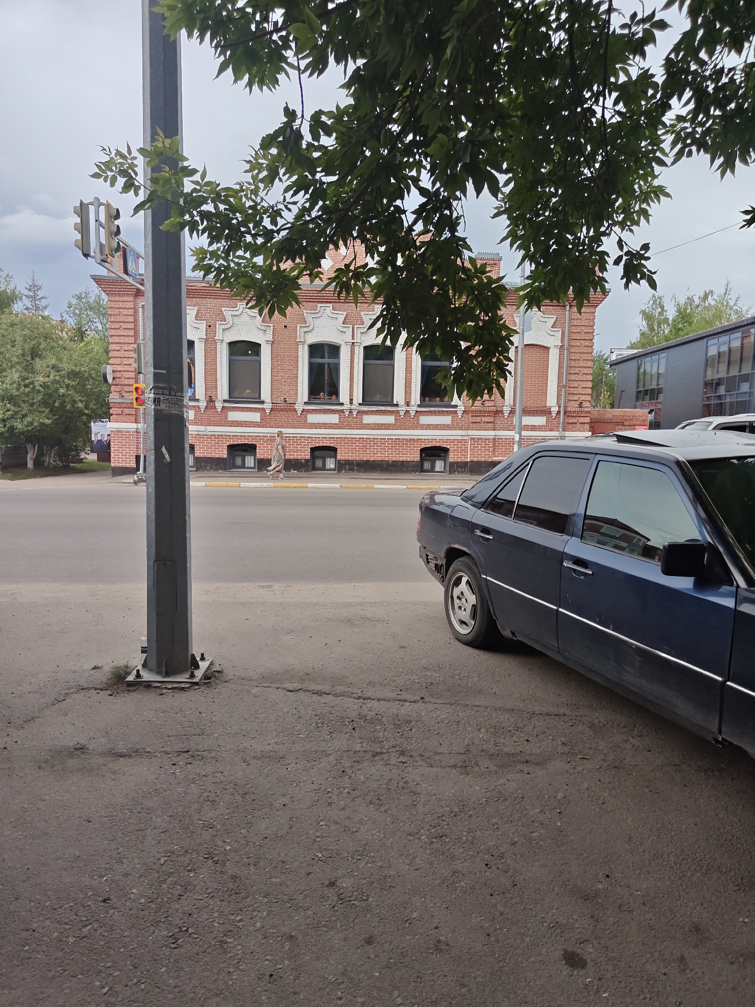 In the foreground, a weathered gray metal lamppost stands on a concrete sidewalk adjacent to an asphalt road. Partially visible in the right side of the frame is the rear of a dark blue sedan with a sunroof and chrome trim. The car's rear bumper appears to be missing or damaged. The road has a yellow dividing line. In the background, a three-story brick building with white decorative trim and arched windows is visible. Several trees with green foliage are scattered throughout the scene. A traffic light with red and yellow lights is mounted on the lamppost. A person is walking on the sidewalk in front of the brick building. The sky is overcast. The image appears to be taken in Petropavl, Kazakhstan.