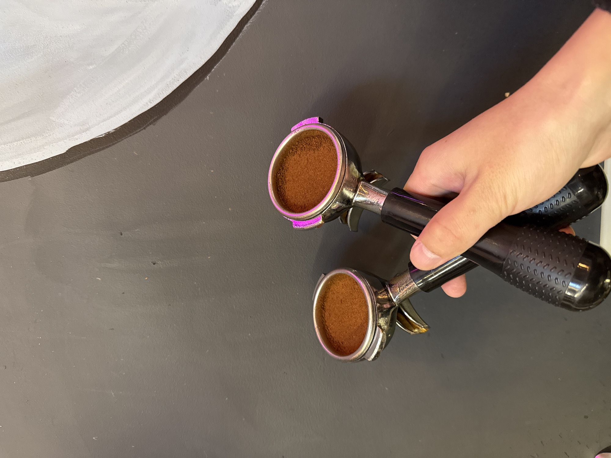 A hand holds two espresso portafilters filled with ground coffee on a gray countertop. The portafilter on top has a purple gasket. A white object is visible in the upper left corner. The location in the city of Moscow, Russia, is not identifiable.