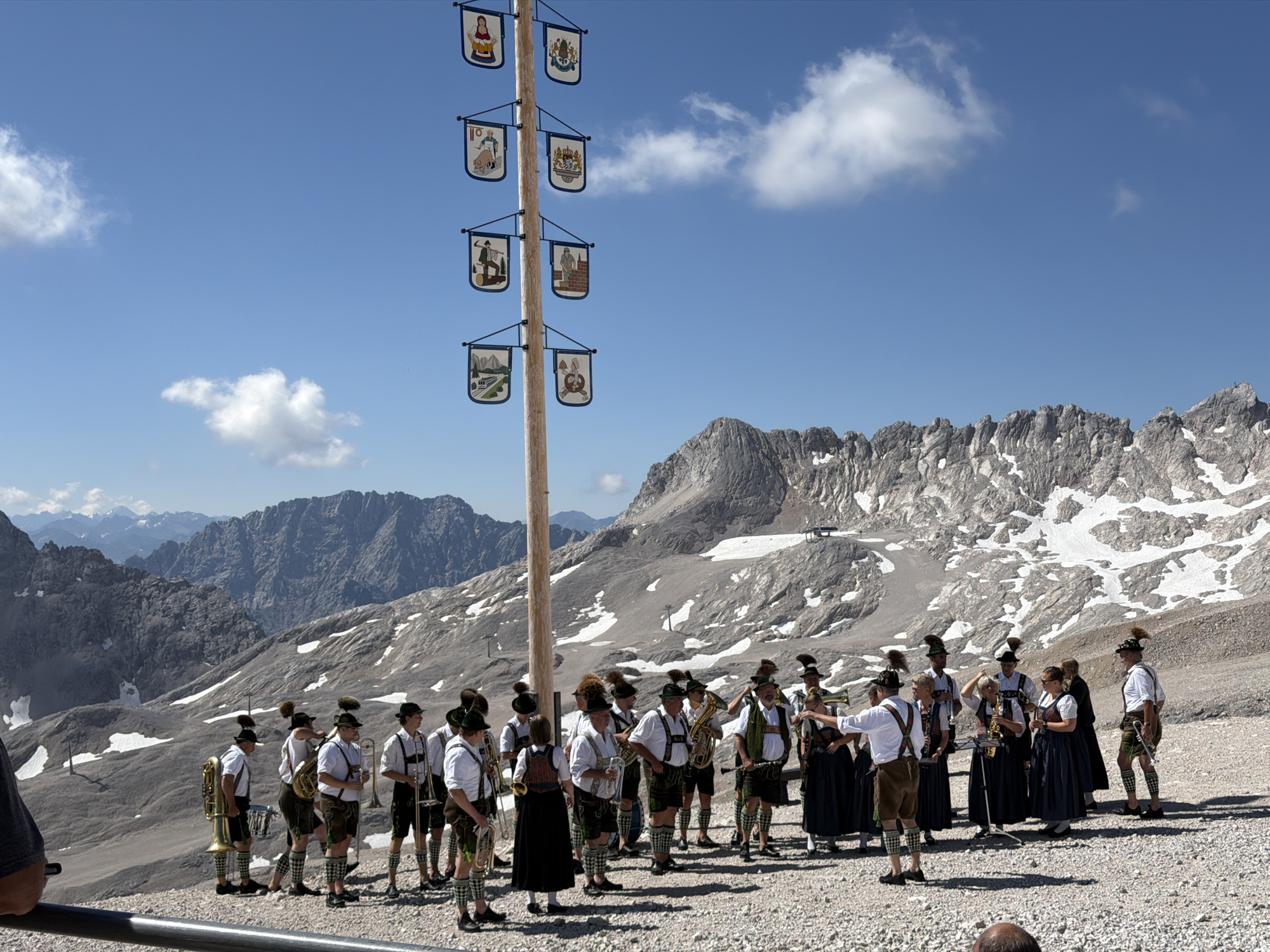 In a mountainous, outdoor environment, a group of approximately 15 individuals, dressed in traditional Bavarian attire, are gathered around a tall wooden pole decorated with various banners. The pole has several levels, with each level holding two banners depicting different scenes or symbols. The individuals are holding musical instruments, including trumpets, tubas, and a drum. Some are standing and looking towards a central point, while others appear to be talking or interacting. The background features a rocky mountain landscape with patches of snow under a clear blue sky with scattered clouds. The ground is covered in gravel. This scene is likely taking place in the city of Leutasch, Austria, known for its alpine scenery and cultural traditions.