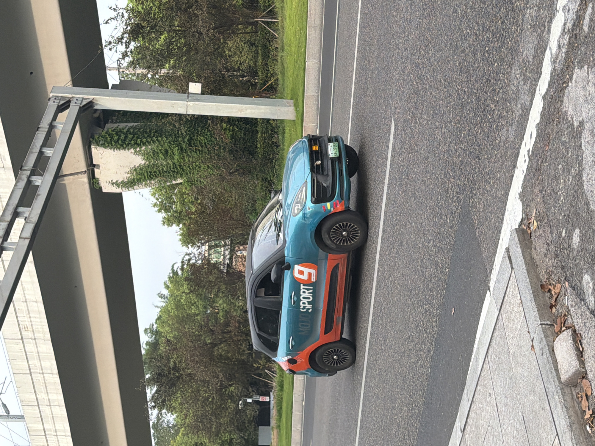 A teal-colored compact SUV with orange and white branding is driving on a paved road in Jiangjin, China. The branding on the side of the car reads "MOJO SPORT" with a large orange number "9". The car has a black roof and black rims. In the background, an elevated concrete overpass runs horizontally across the top of the image, supported by large concrete pillars. Lush green trees and vegetation line the sides of the road and grow around the base of the pillars. The sky is overcast. A grey concrete sidewalk with fallen leaves is visible in the foreground. The license plate of the car is visible and reads "BXL29888".