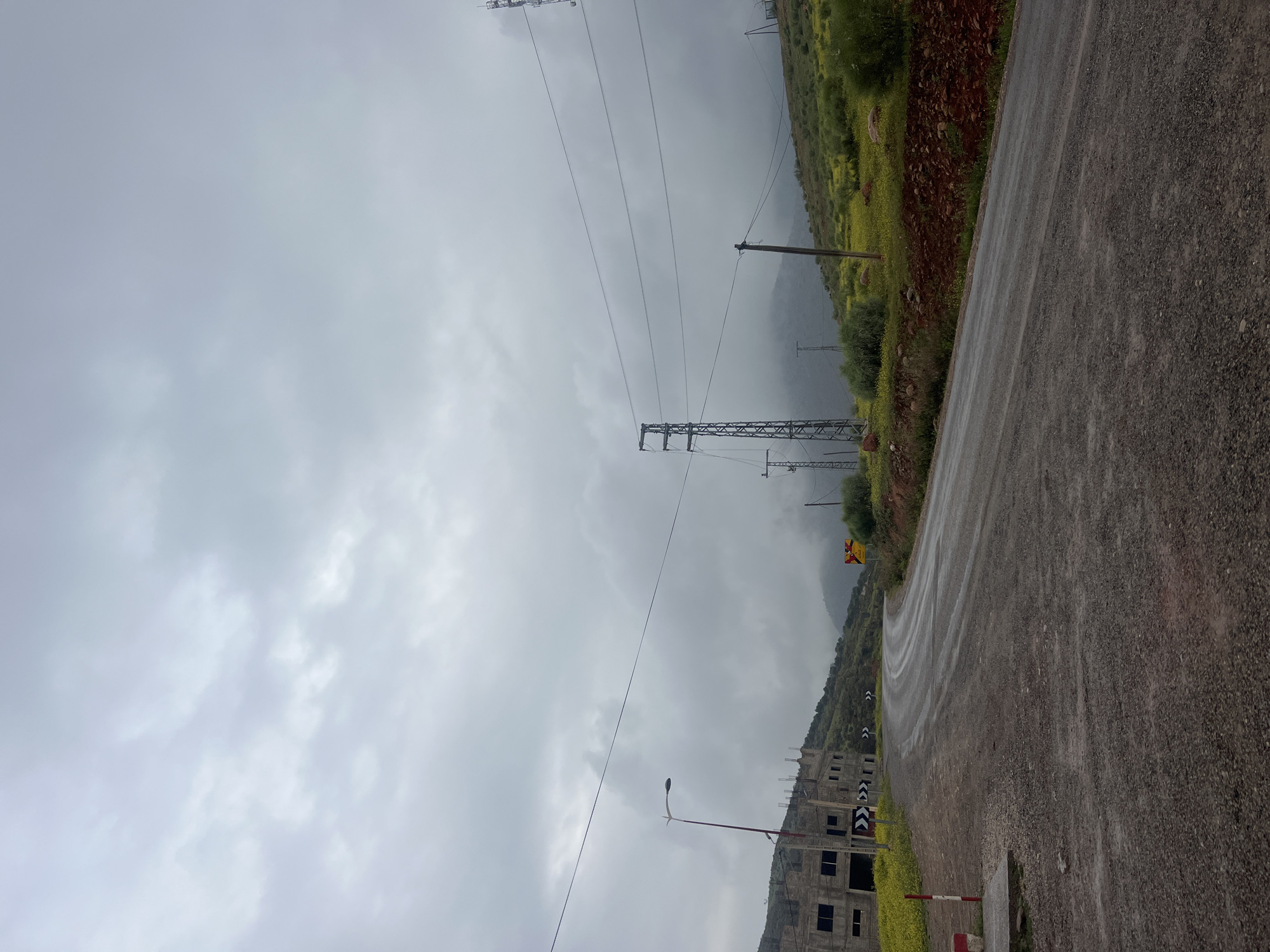 A cloudy, rainy day in Ouaouizert, Morocco. The image is taken on a wet, asphalt road winding up a mountainside. Vegetation, including green bushes and small yellow flowers, lines the side of the road. An unfinished, multi-story concrete building is visible on the lower side of the road. Several utility poles and electrical wires stretch across the sky. Roadside markers, black and white chevrons, are present along the edge of the road. A yellow warning sign is visible higher up the road. No people are visible.