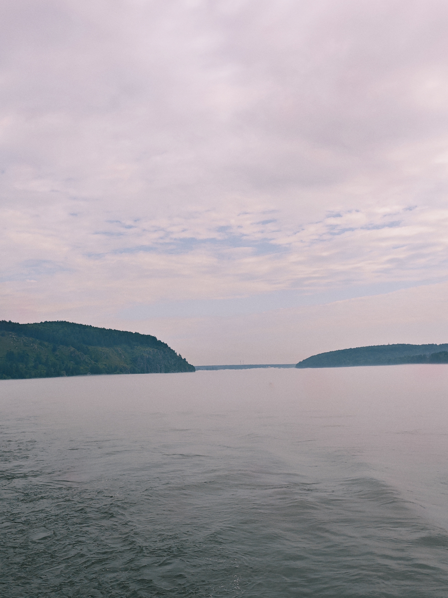 A wide-angle, low-angle shot captures a serene river scene under a pale, hazy sky. The water in the foreground is choppy, with visible ripples and small waves. In the distance, rolling green hills dotted with trees rise from the riverbank. The sky is filled with soft, diffused clouds, casting a gentle, muted light over the landscape. The overall impression is one of tranquility and natural beauty. This scene is located in Atamanovo, Russia, likely along the Yenisei River.