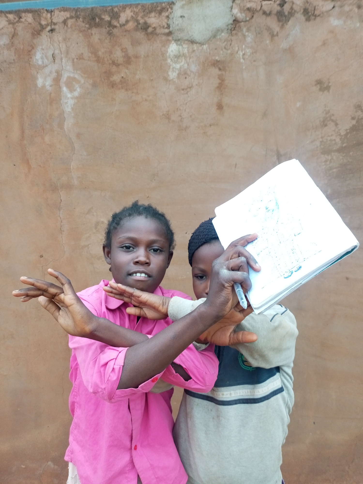 Two children are pictured in front of a mottled brown wall. The child on the left is a girl wearing a bright pink collared shirt with red buttons and has her arms crossed in front of her chest. The child on the right is wearing a light gray sweatshirt with a dark blue and light blue striped accent and a black beanie. This child is holding a white notebook and a pen. The wall behind them appears to be made of concrete with visible cracks and discoloration.