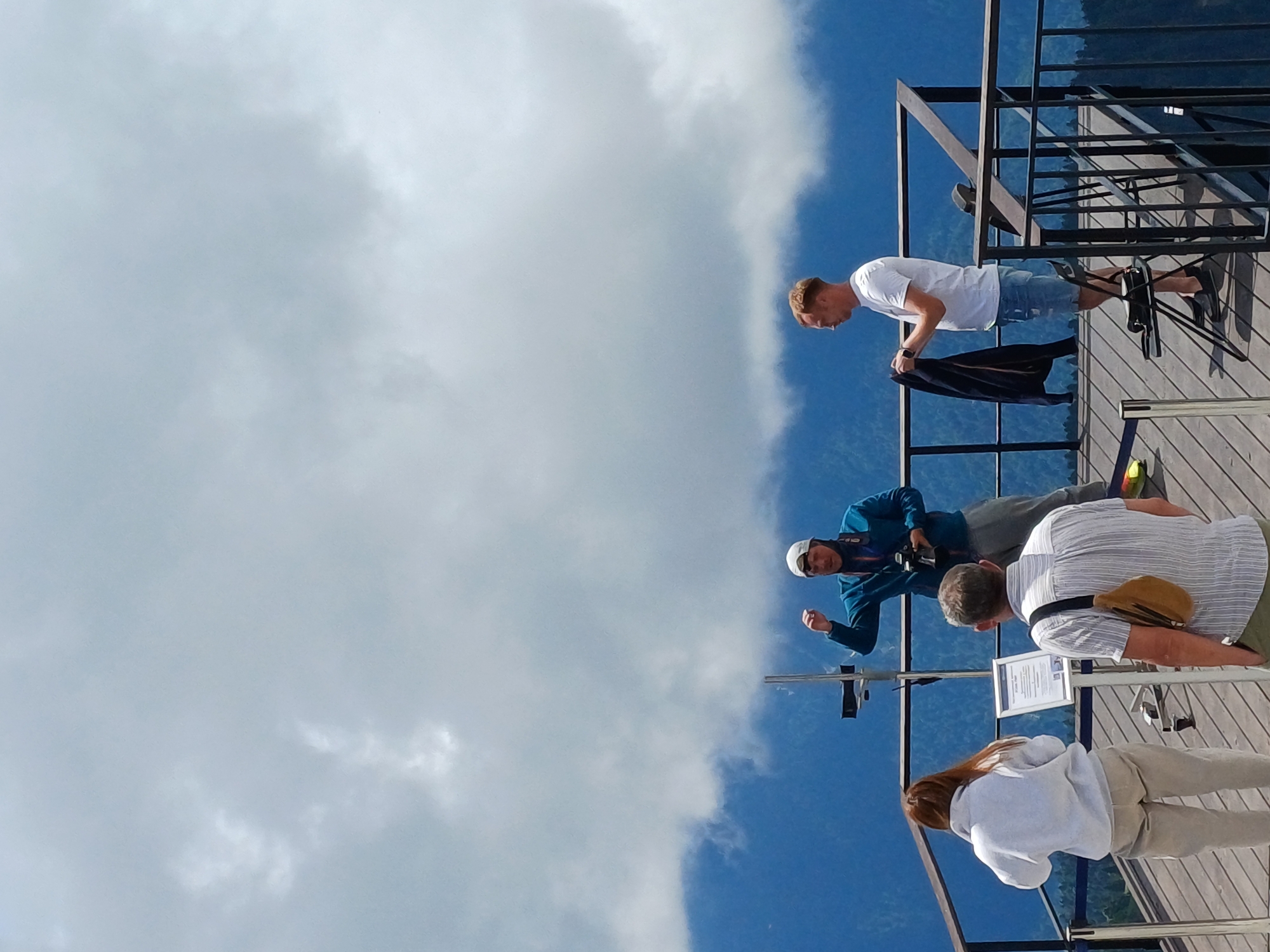 This image depicts a group of people on an observation deck in Estosadok, Russia, with a backdrop of mountains and clouds. In the foreground, a woman with red hair wearing a light gray hoodie and beige pants faces away from the camera. To her right, a man in a striped shirt and khaki crossbody bag also faces away. In the mid-ground, a man in a blue jacket and a white baseball cap is holding a camera and gesturing with his left hand. To the far right, a man in a white t-shirt and denim shorts stands looking to his left, holding a dark jacket. The observation deck is constructed of wooden planks and metal railings. The background shows lush green mountains partially obscured by thick white clouds.