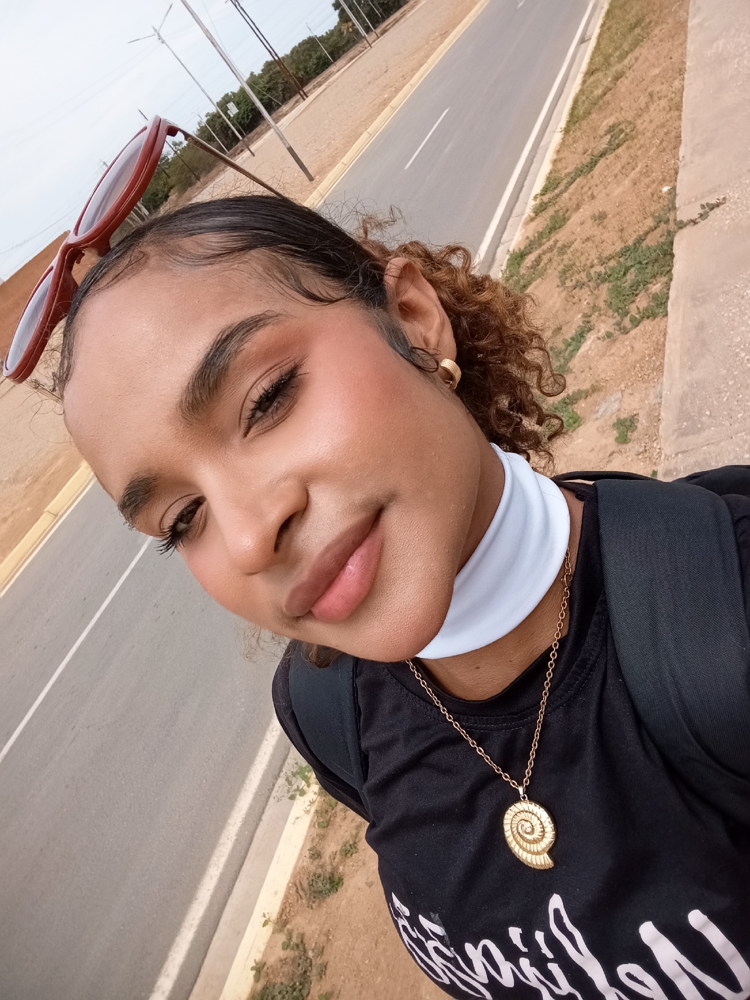 A young woman with warm skin tones is featured in a close-up selfie, taken outdoors. Her dark, curly hair is styled with subtle baby hairs around her forehead, and reddish-brown sunglasses rest on top of her head. She wears light makeup, including defined eyebrows, long eyelashes, blush on her cheeks, and glossy lips, and she looks directly at the camera with a gentle smile.
Her accessories include a gold hoop earring in her right ear, a white neck gaiter or band, and a gold chain necklace with a prominent spiral shell-shaped pendant. She is wearing a black t-shirt that has visible white text on the front, appearing to be a stylized word such as "familiar." A black backpack strap is visible over her right shoulder.
The background shows a paved road with a solid white line, suggesting a main road or highway. To the right of the road is a dirt and gravel shoulder with sparse green vegetation and a concrete barrier. In the distance, utility poles and trees are visible under an overcast or hazy sky. The image was captured during the daytime. The scene suggests a casual moment, possibly taken while on a journey or stopped by the roadside. The location context provided is Porlamar, Venezuela.