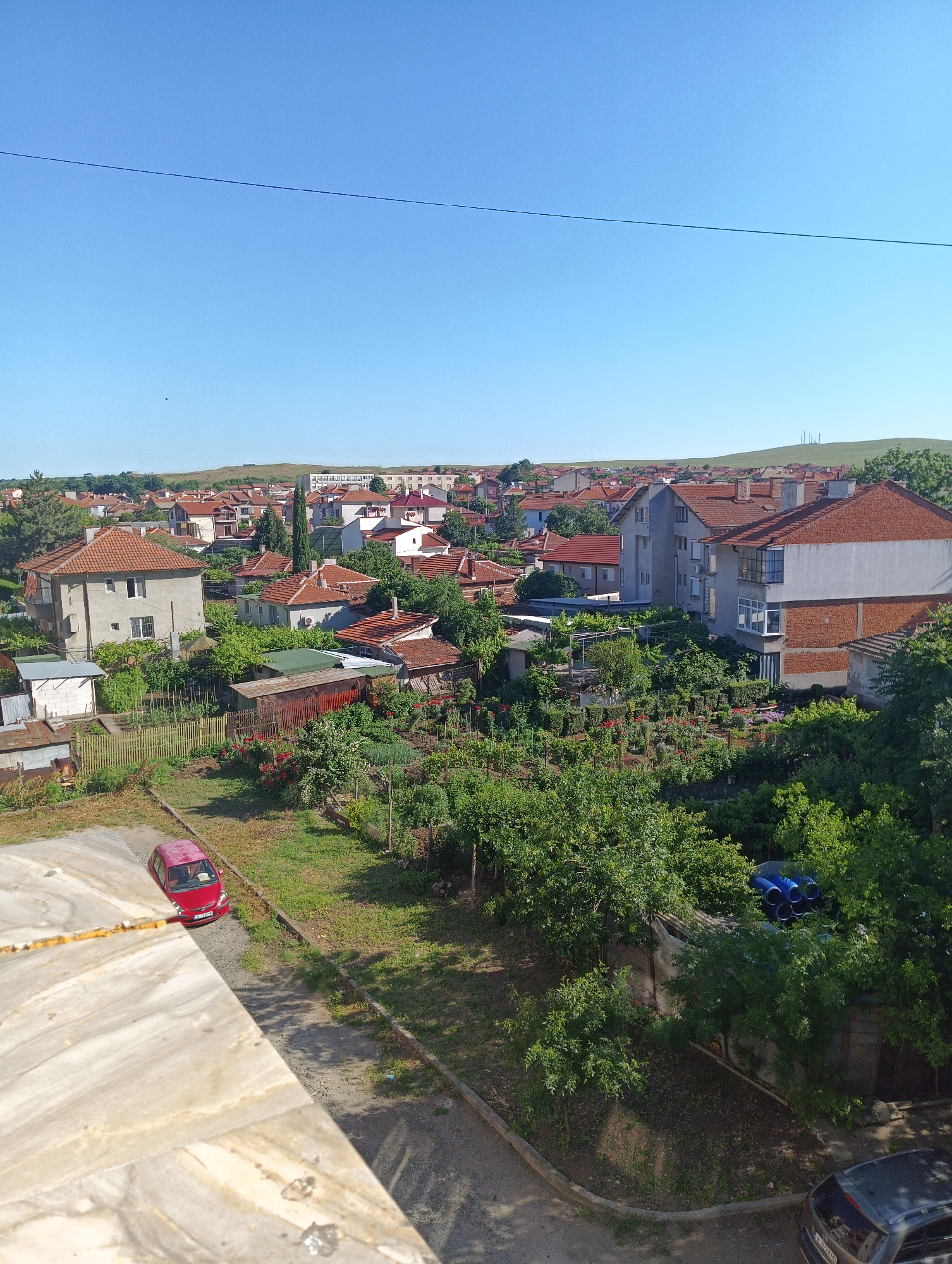This is an exterior, medium-shot, high-angle photograph of a residential area in Karnobat, Bulgaria, on a clear, sunny day. The foreground features a light-colored, textured surface, possibly a balcony railing, angled from the lower left to the mid-right. Below this railing, a narrow, paved area runs diagonally, occupied by a red, four-door hatchback car parked. The car's license plate is partially visible, showing text and numbers.
Beyond the paved area, a garden with dense green foliage, including trees and shrubs, stretches across the middle ground. Within the garden, several low-rise buildings with red-tiled roofs are visible, some appearing to be homes and others outbuildings or sheds. A weathered wooden fence runs through the garden. Further into the distance, a densely packed residential neighborhood with houses and apartment buildings extends to the horizon. These buildings also feature red-tiled roofs. The background consists of rolling green hills under a clear blue sky. A power line stretches horizontally across the upper portion of the sky. There are no people visible in the image.