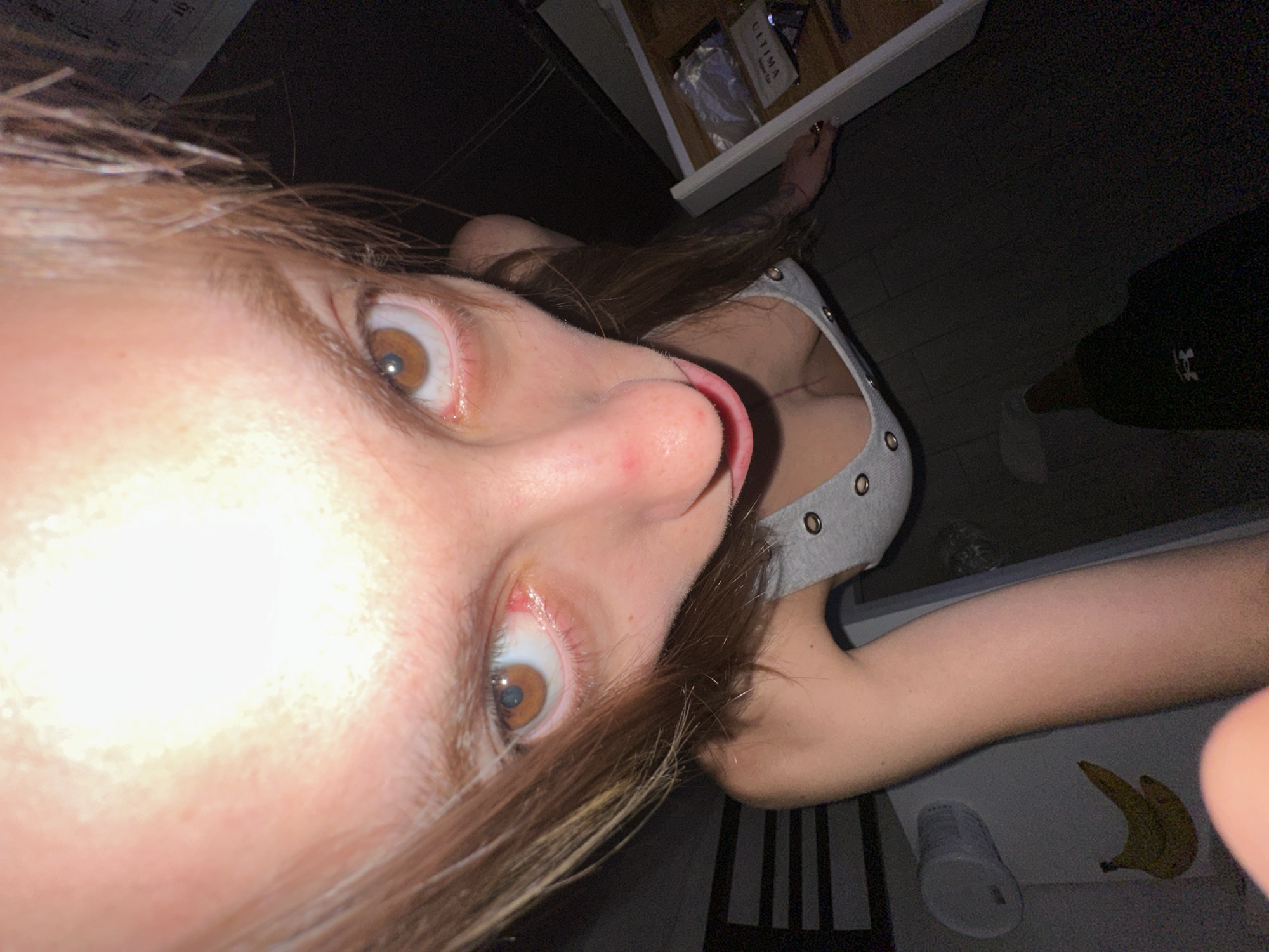 The media file shows a close-up, low-angle, slightly blurred image of a person's face and upper body. The person has light brown hair that is falling over their face, obscuring part of their left eye. Their right eye is clearly visible, with a brown iris. Their mouth is slightly open, with their tongue peeking out. They are wearing a gray tank top with grommet details. The background is dark, with some objects visible, including what appears to be a drawer or shelf with items inside, a white cup, and a banana. The lighting is dim, suggesting it might be nighttime or indoors with low light. The overall impression is a candid, playful, or perhaps mischievous moment captured. There is some text visible on an item in the drawer, reading "ULTIMA." The scene appears to be indoors, possibly in a home environment, given the presence of everyday objects. The image is shot from a perspective that emphasizes the person's face and their interaction with the camera.