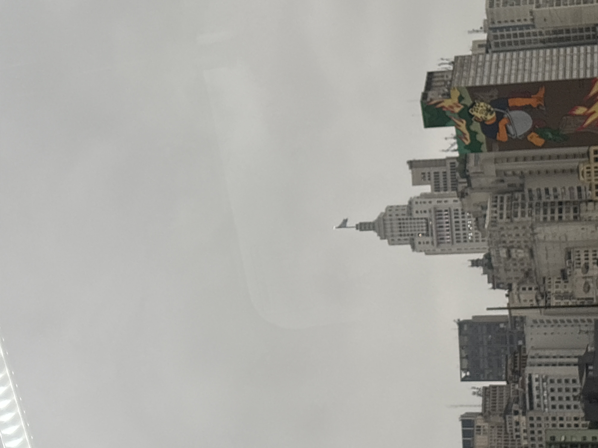 This image captures a cityscape in São Paulo, Brazil, likely in the central business district, given the presence of numerous skyscrapers. A tall, distinctive building with a flag at its apex stands out in the center, characteristic of an Art Deco style, possibly the Edifício Altino Arantes (Banespa Building). Surrounding it are various other buildings of different heights and architectural styles, some appearing older and others more modern.
On the right side of the image, a large mural adorns the side of a building. The mural depicts a stylized figure with a mask, possibly a character from mythology or folklore, surrounded by elements resembling flames or abstract shapes in green and red. The figure appears to be holding a gray bucket.
The sky is overcast, suggesting a cloudy or possibly rainy day, which contributes to the muted tones of the cityscape. The overall impression is that of a dense urban environment, a common sight in the heart of São Paulo. There are no visible people or immediate human activity in the frame, focusing solely on the architecture and the prominent mural. The perspective is from a slightly elevated viewpoint, looking across the city.