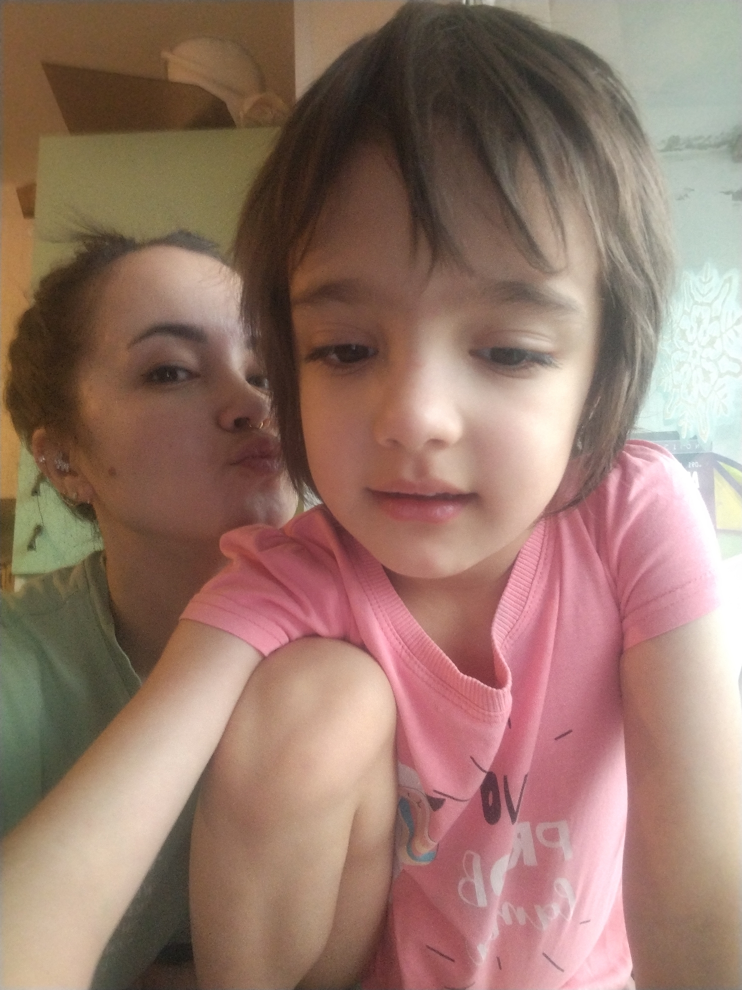 Two individuals are in an interior setting, possibly a residence. A child is positioned in the foreground, wearing a pink short-sleeved shirt with a graphic design. The child has short brown hair and appears to be looking downward. Behind the child, a woman is visible in the background, making a kissing face towards the camera. The woman has dark hair pulled back into a bun and is wearing a light green shirt. The background includes indistinct elements of a room interior, with a partial view of a green wall, a shelf with objects, and possibly patterned wallpaper. The image appears to have been taken indoors in Noviy, Russia.