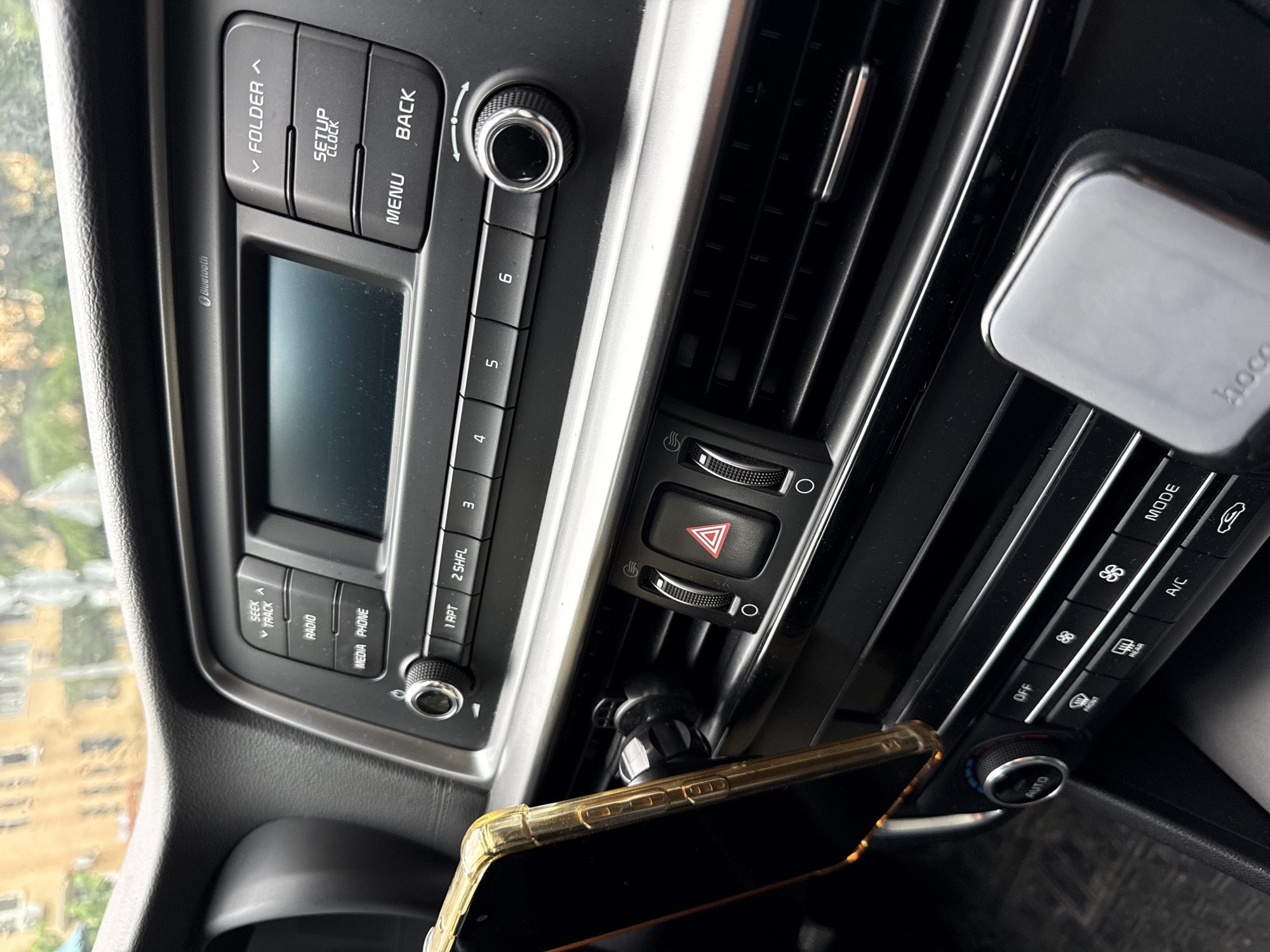 The image shows the interior of a car, specifically the center console area. There is a car stereo unit with buttons labeled "FOLDER," "SETUP," "BACK," "MENU," and numbered buttons. Below that are buttons labeled "SEEK," "RADIO," "MEDIA," and "PHONE." An air vent is visible, with hazard light button and temperature control dial. A smartphone in a gold case is mounted on the air vent, and a "hoco" brand device is also visible. The location is not identifiable as Sochi, Russia from the image alone.