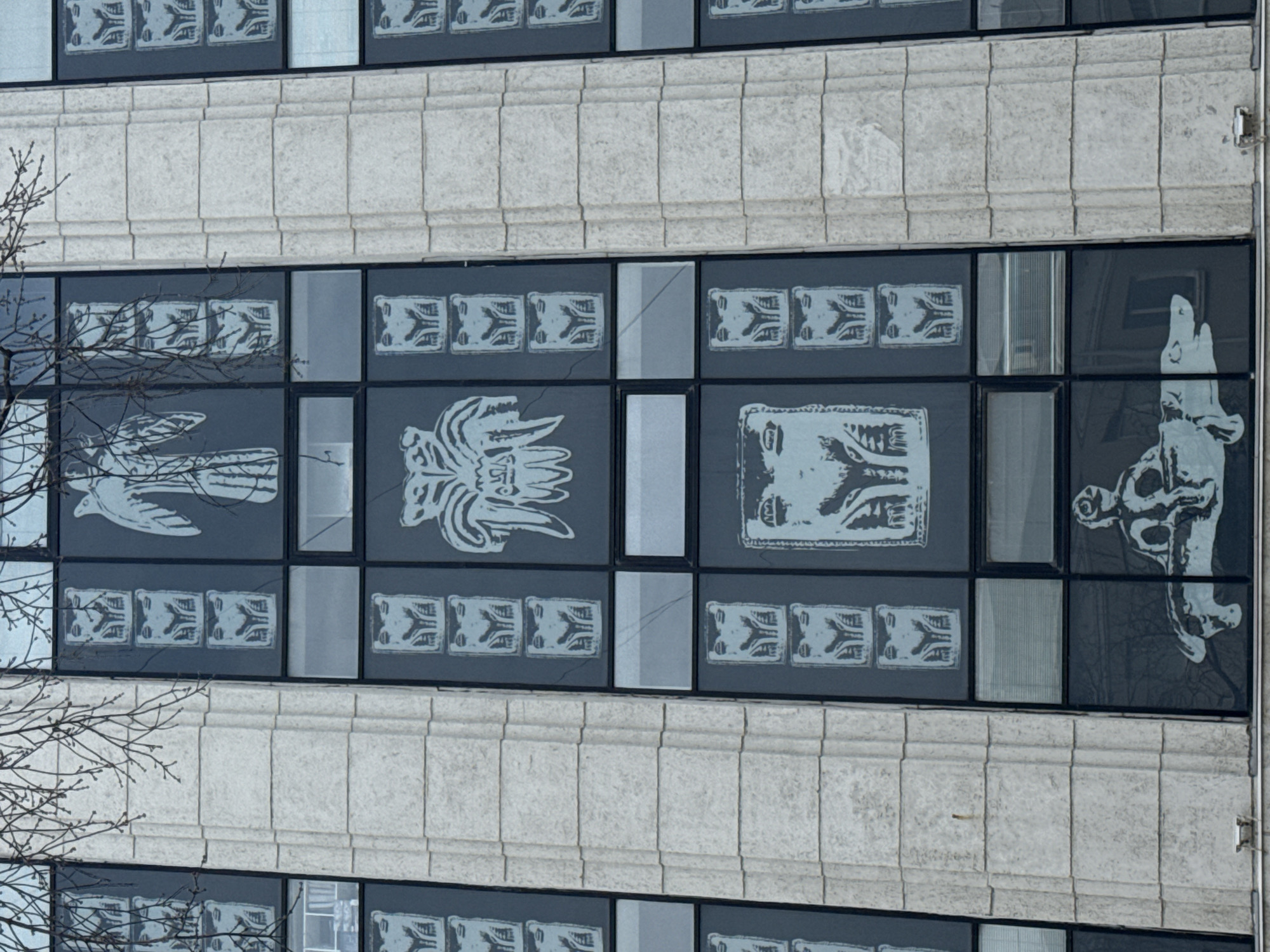 The image shows a section of the "Legend of Perm Bear" building facade in Perm, Russia. The facade is constructed of light-colored stone blocks arranged in horizontal rows. A row of windows is visible between stone rows, each adorned with grayscale illustrations or patterns. From left to right, these include a bird, decorative patterns, a complex design, a portrait of a face, and a hand holding what appears to be a skull. The building's reflective windows show a partial view of the surrounding bare tree branches and other buildings across the street.