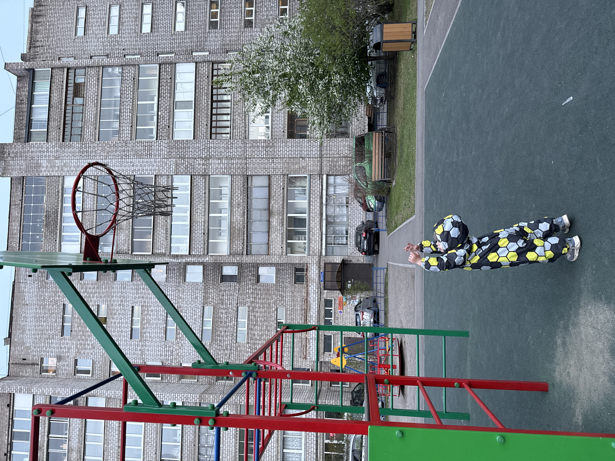A daytime shot shows a child in a black and yellow honeycomb-patterned jumpsuit standing on a green surface outdoors, holding a ball overhead with both hands. In the background is a tall, multi-story brick building with rows of windows. To the left of the child is a basketball hoop and a playground structure with green and red metal bars. Further back, trees and a parked black car are visible. The scene appears to be in Krasnoyarsk, Russia.
