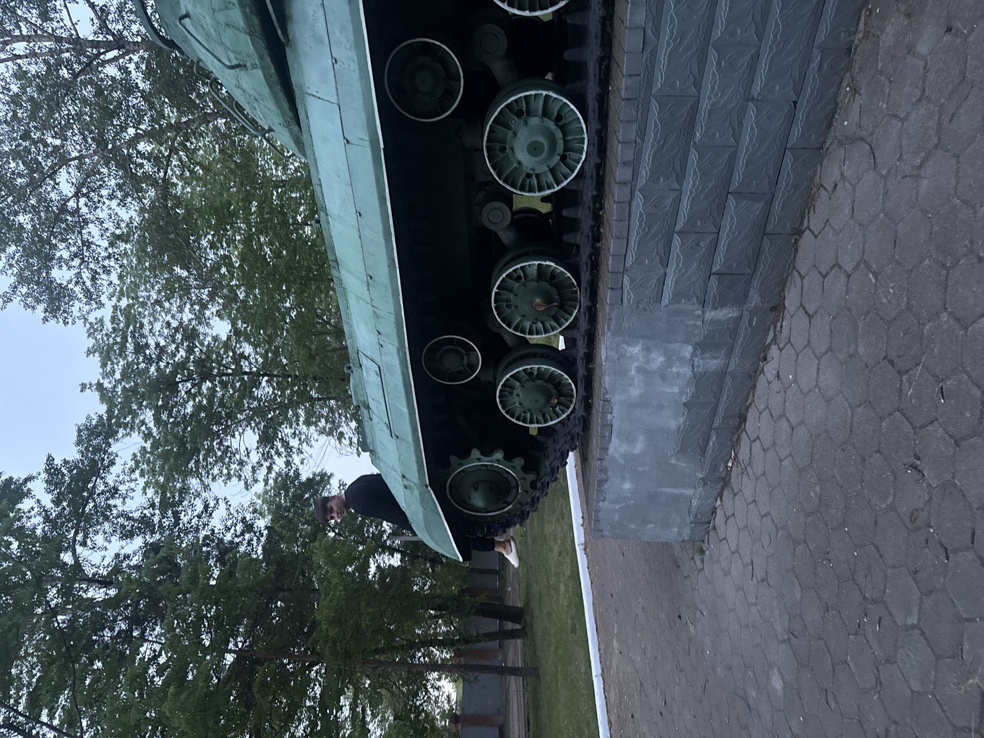 The image captures an outdoor scene featuring a military vehicle displayed as a monument, with a person standing alongside it.
The main subject is a large, green military vehicle, resembling an armored personnel carrier or infantry fighting vehicle, mounted on a dark grey stone pedestal. The vehicle's tracks and multiple road wheels are visible, with the wheels also painted green, some accented with white trim. The stone pedestal is composed of rectangular blocks adorned with a textured, decorative pattern.
A young man, dressed in a dark long-sleeved shirt, dark pants, and white shoes, is partially visible standing to the left of the military vehicle, looking towards the viewer. His posture suggests he is observing or posing with the monument.
The ground surrounding the monument is paved with dark grey stones; some areas feature a hexagonal pattern, while others closer to the pedestal are rectangular. In the background, numerous trees with lush green foliage stand against a bright, overcast sky. The diffused lighting suggests it is daytime, possibly late afternoon or early evening. There is no discernible text visible in the image. The scene depicts a public memorial or historical display, consistent with locations found in Birobidzhan, Russia.